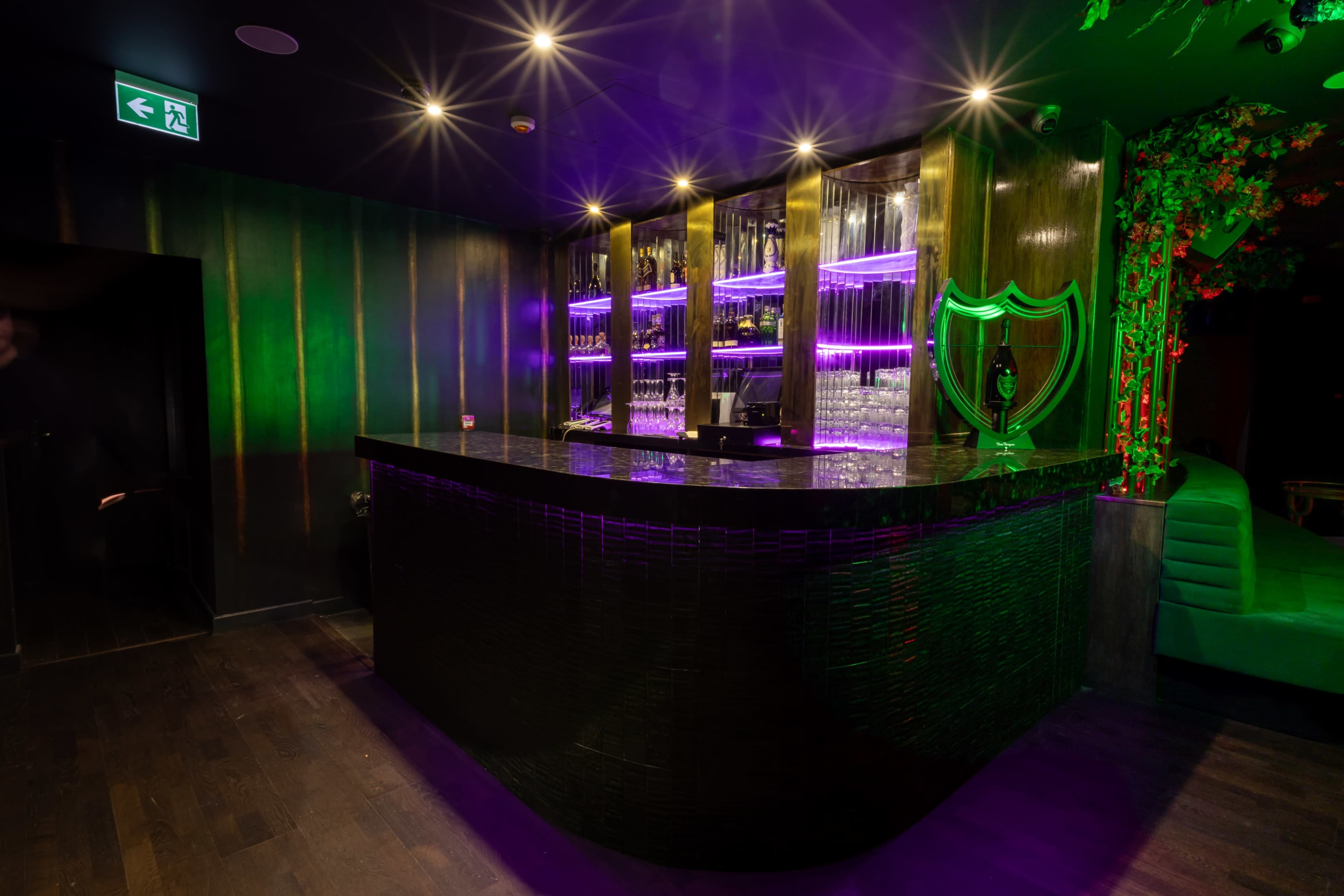 The image depicts a bar area with a sleek dark counter, illuminated shelves filled with glassware, and purple lighting accents.