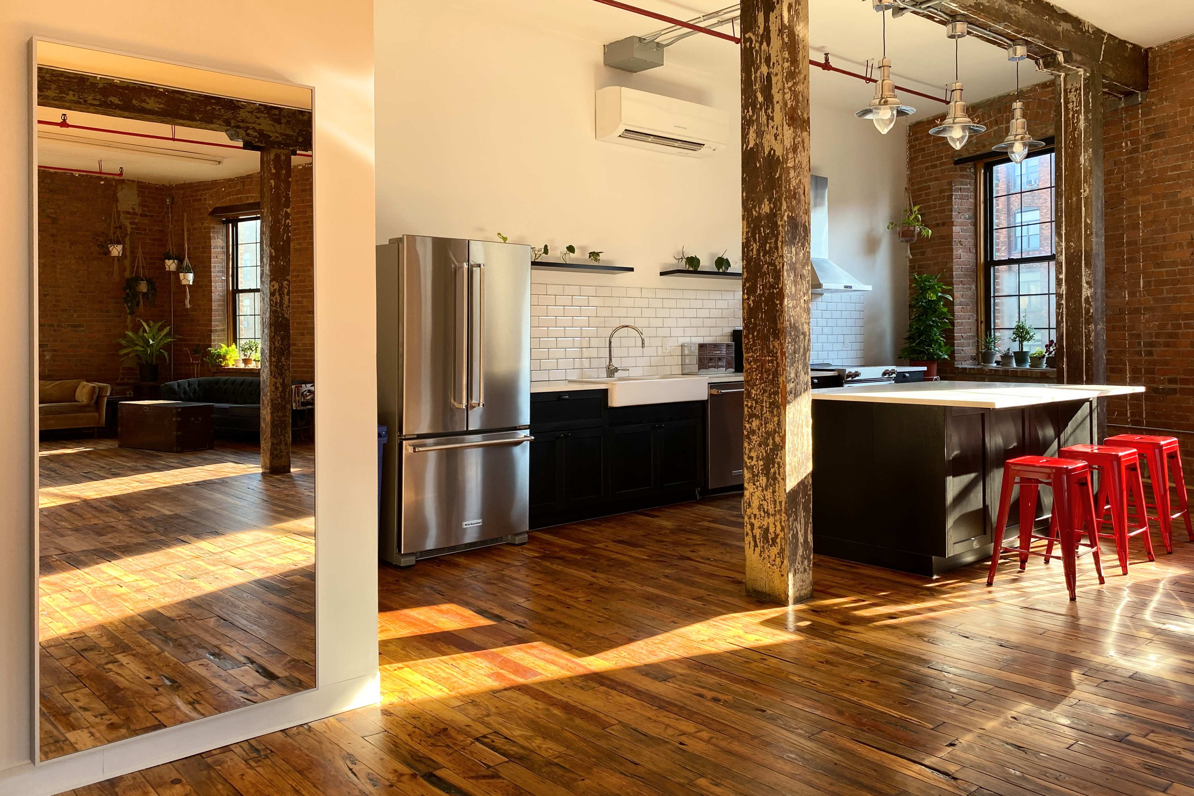 A modern kitchen with exposed brick walls, wooden flooring, stainless steel appliances, and red metal stools positioned at a kitchen island, alongside a large mirror reflecting part of the space.