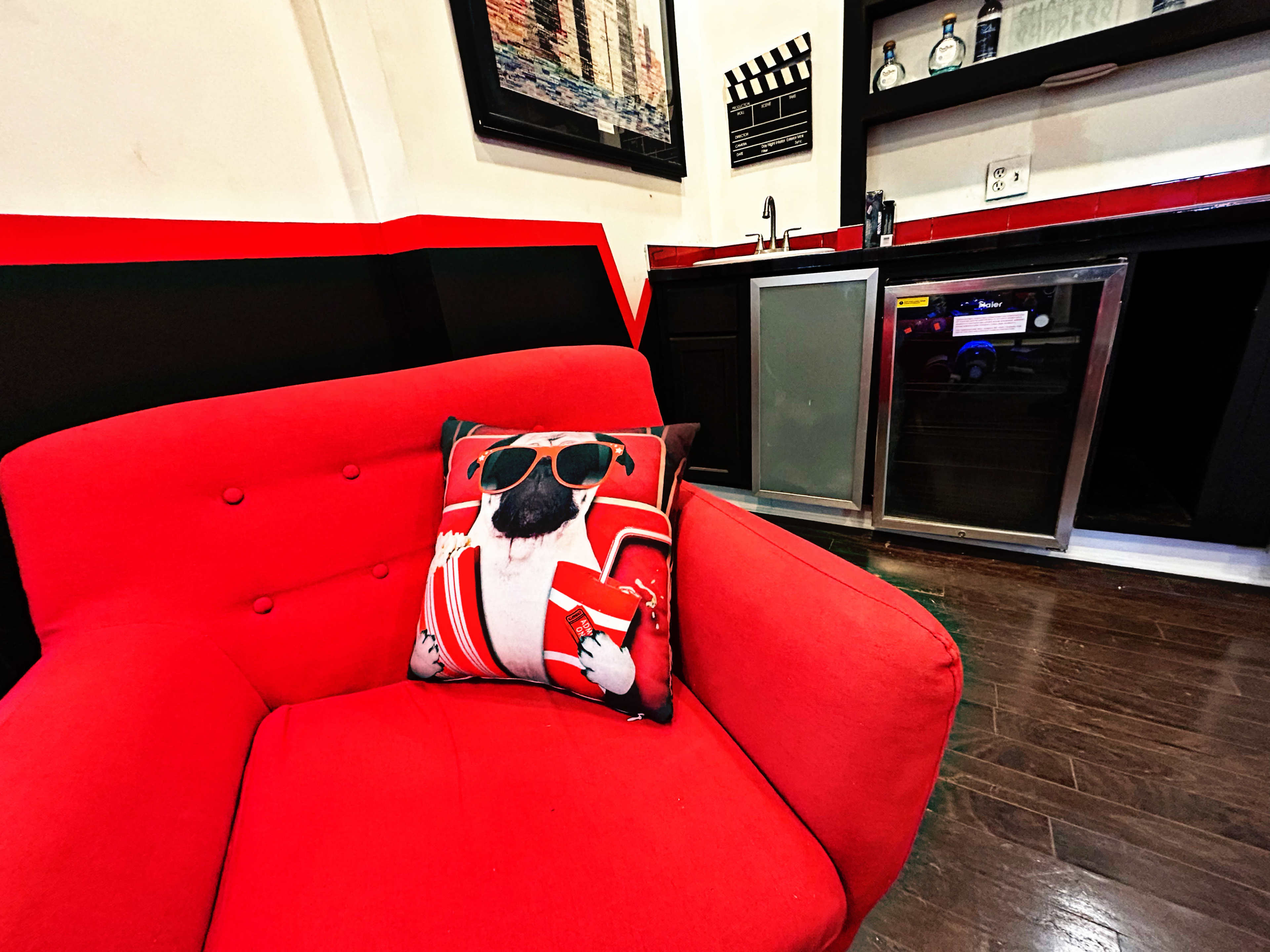 A red armchair with a decorative pillow featuring a dog in sunglasses is positioned near a kitchenette with dark cabinetry and modern appliances.