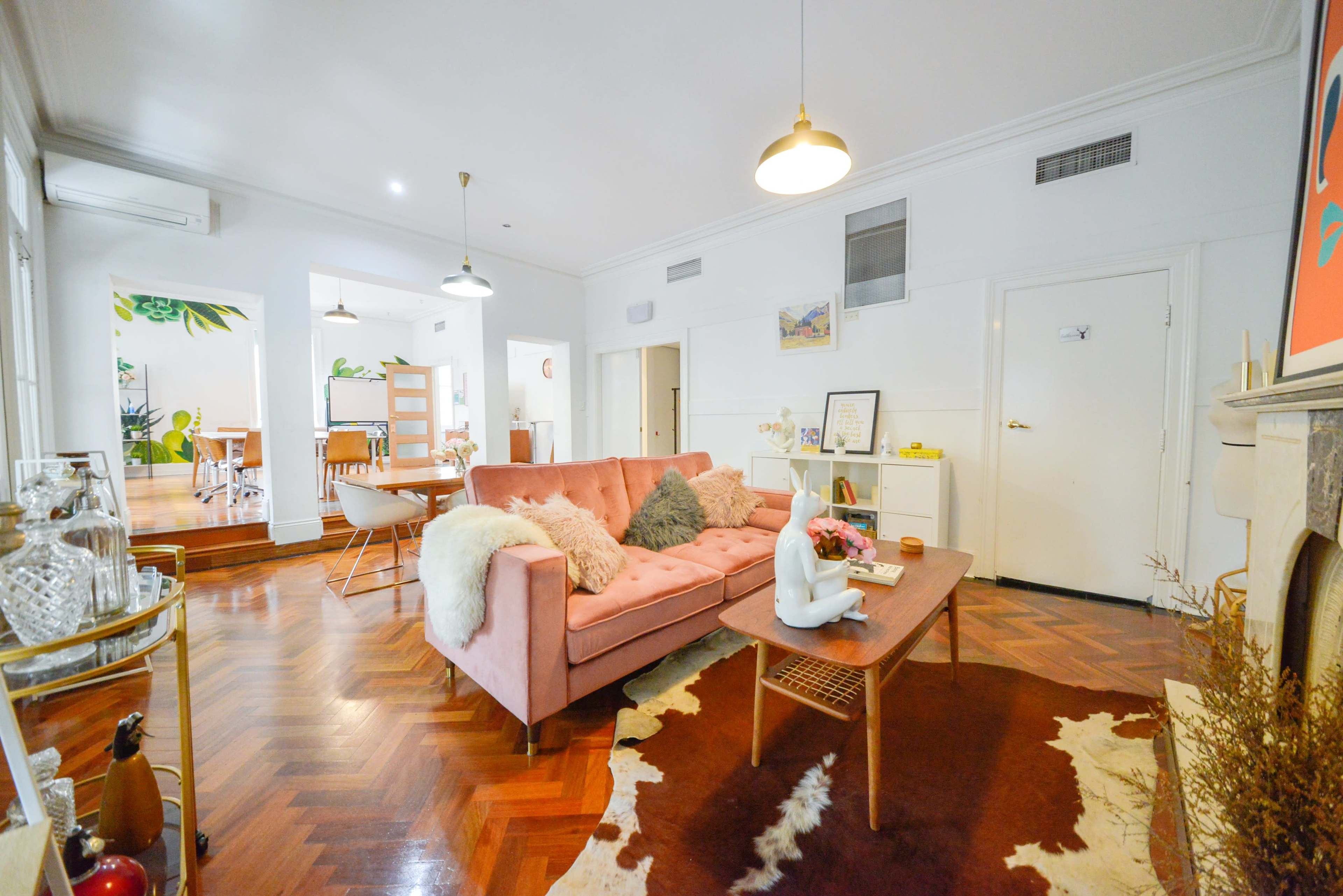 A living room with a pink sofa, wooden coffee table, and a decorative rug, featuring light-colored walls and a plant design in the background.