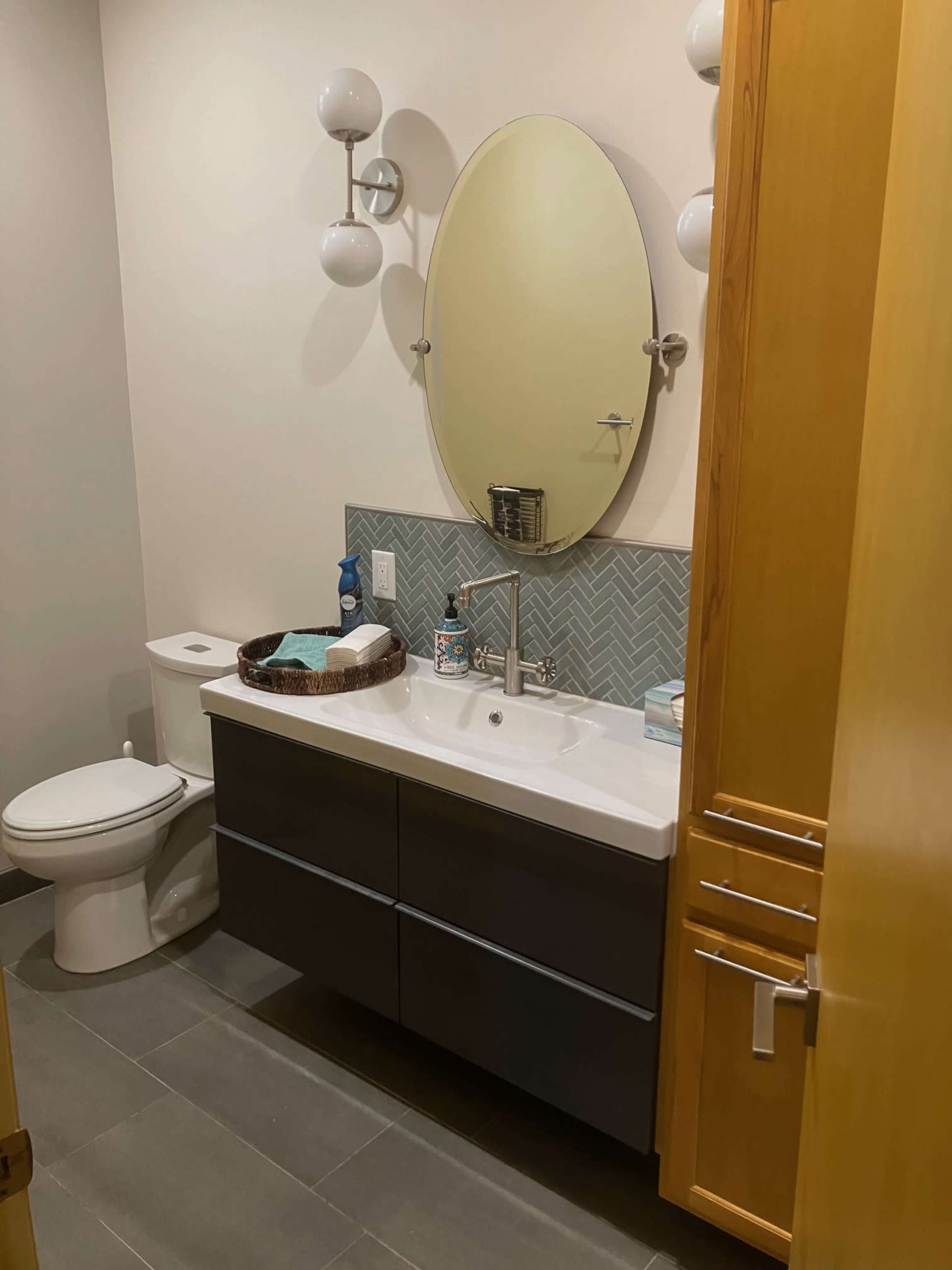 The image shows a modern bathroom featuring a gray vanity with a sink, a round mirror, a toilet, and a herringbone-patterned backsplash.