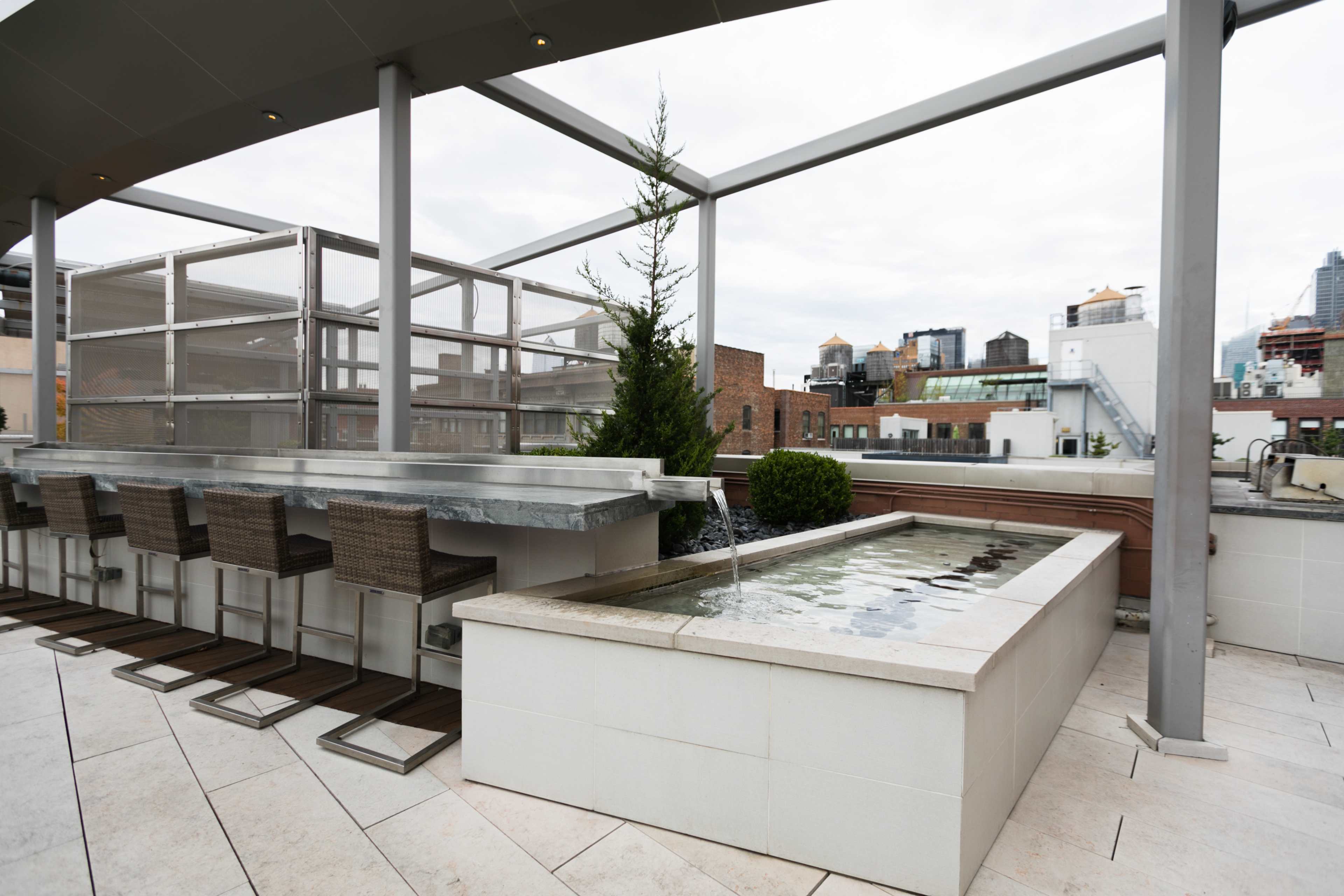 The image shows a modern rooftop terrace featuring a bar with stools, a small water fountain, and city buildings in the background.
