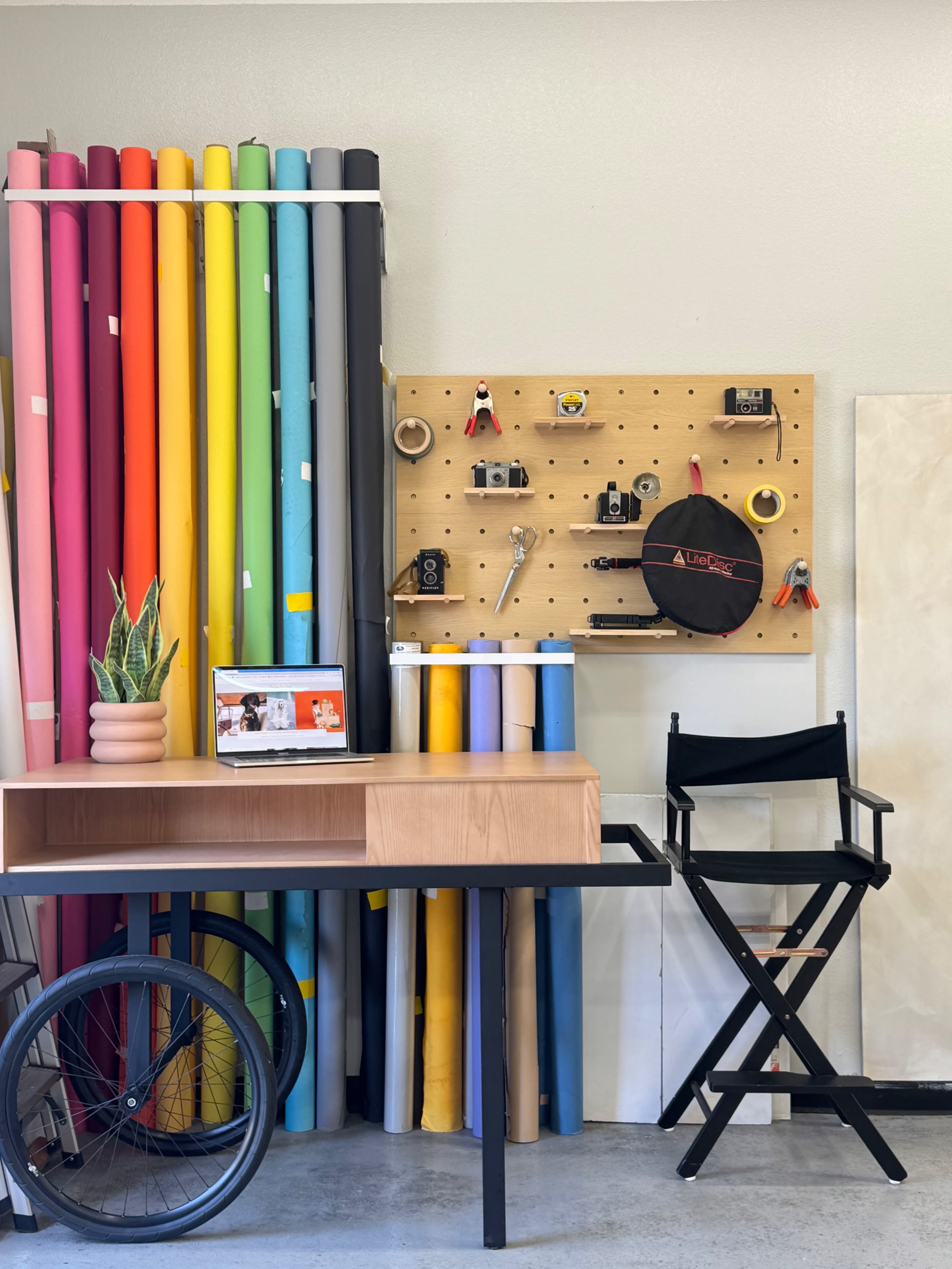 The image shows a workspace featuring rolls of colored paper, a wooden table, a director's chair, and a pegboard with various tools mounted on it.