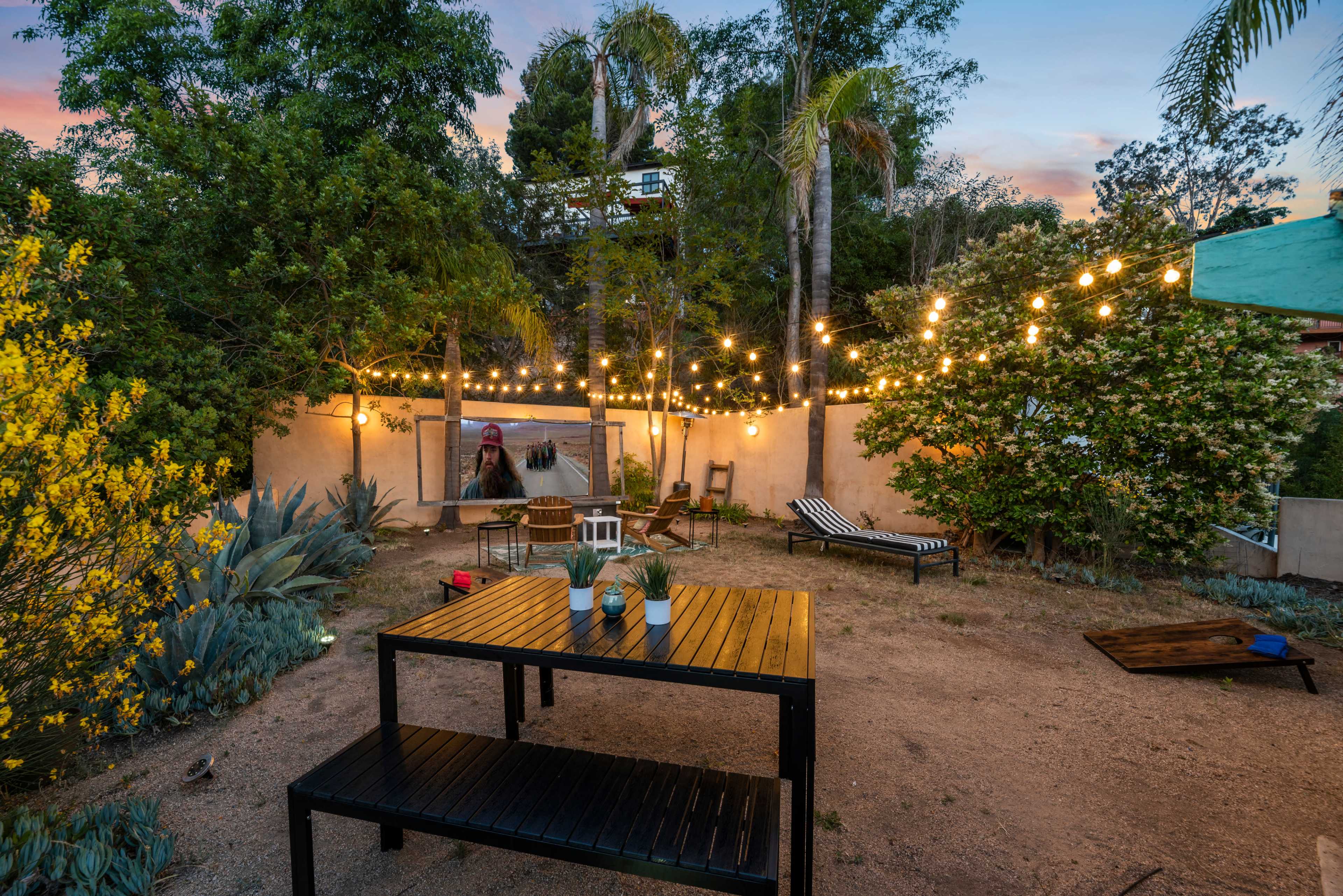 The image shows a landscaped backyard with a wooden table, string lights, and lounge chairs surrounded by greenery and a mural on the wall.