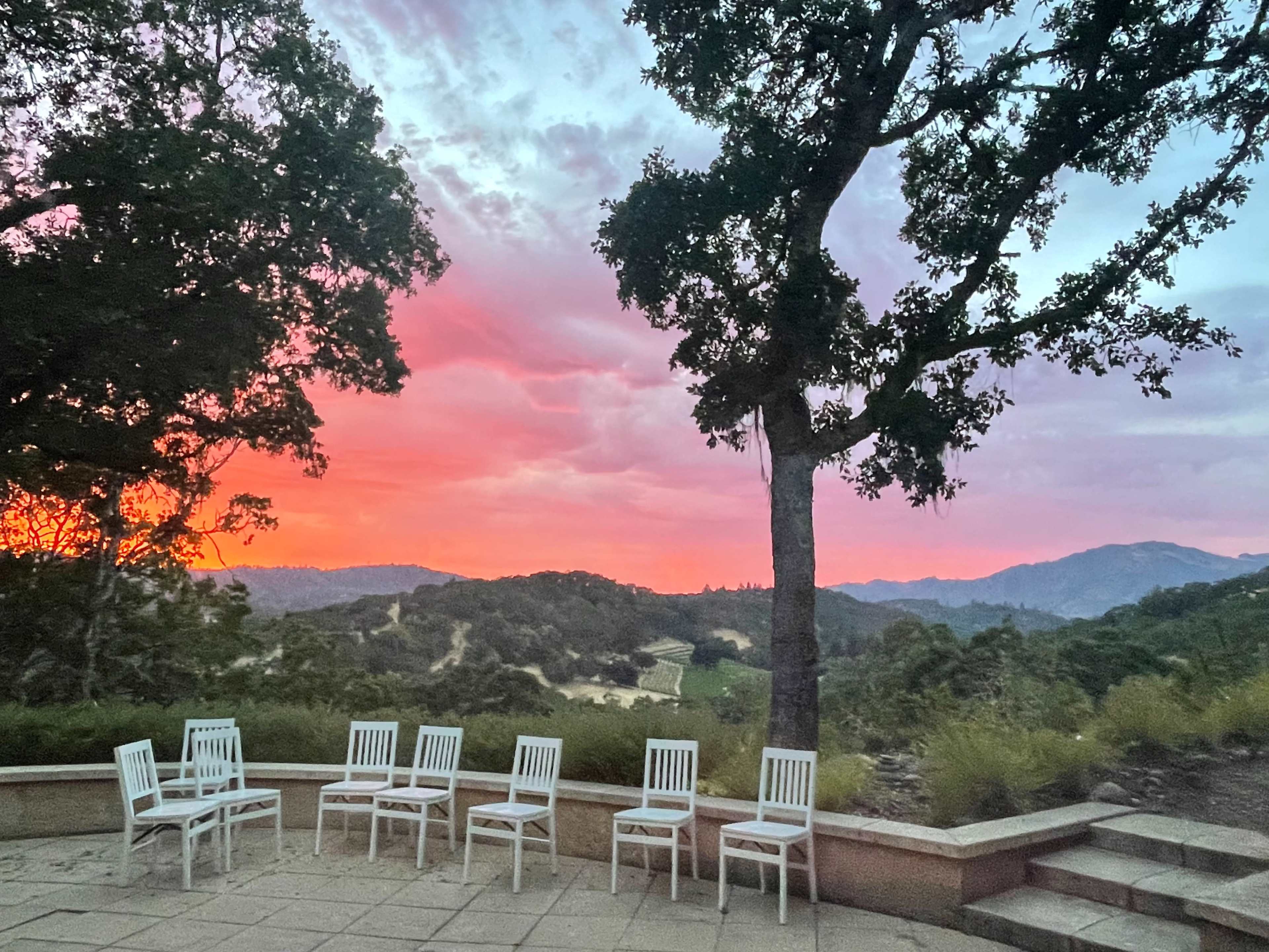 White chairs are arranged in a semicircle on a stone patio overlooking a landscape bathed in colorful sunset hues.