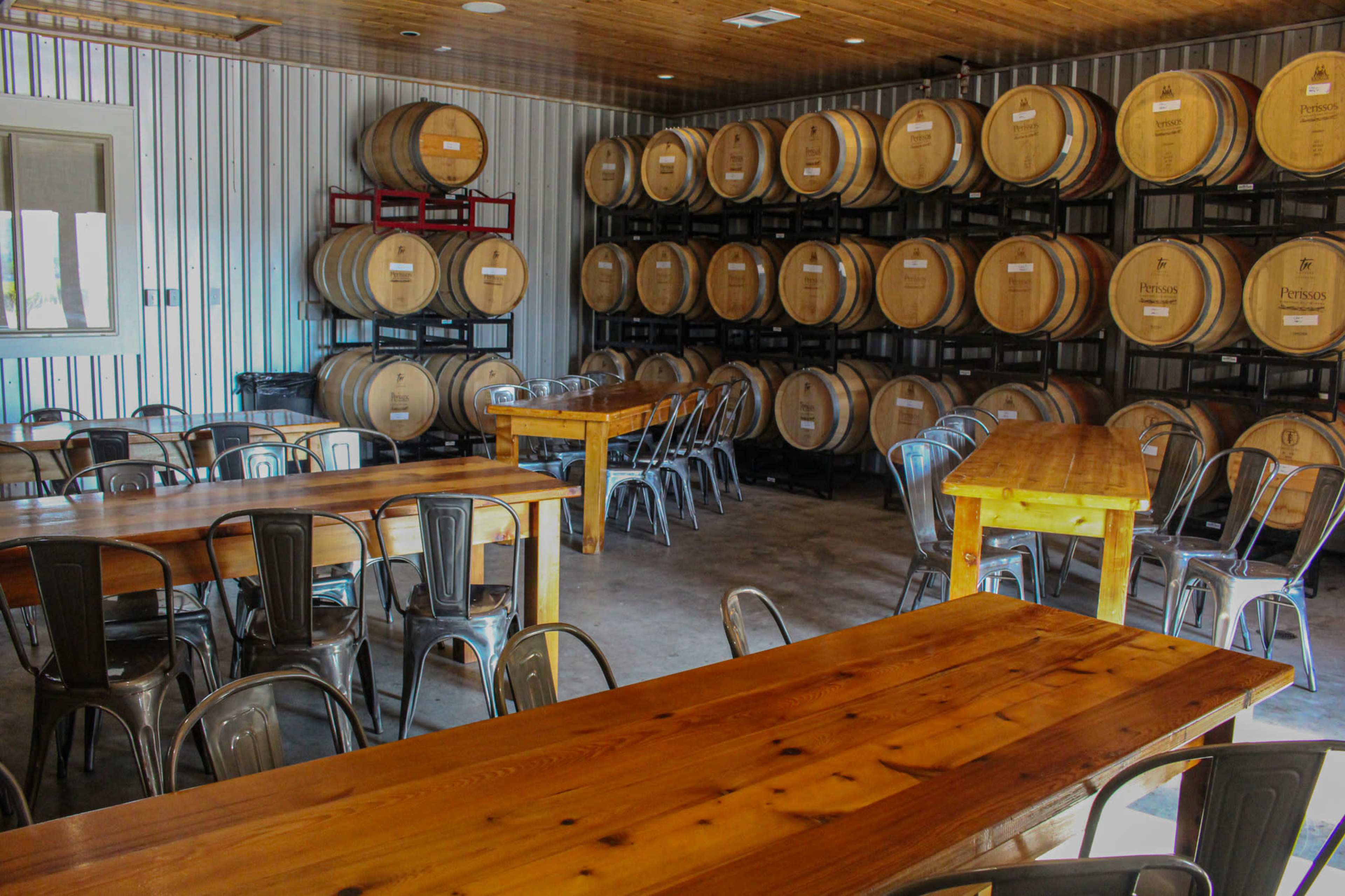 A tasting room features wooden tables and metal chairs arranged among rows of stacked wine barrels against a metallic wall.