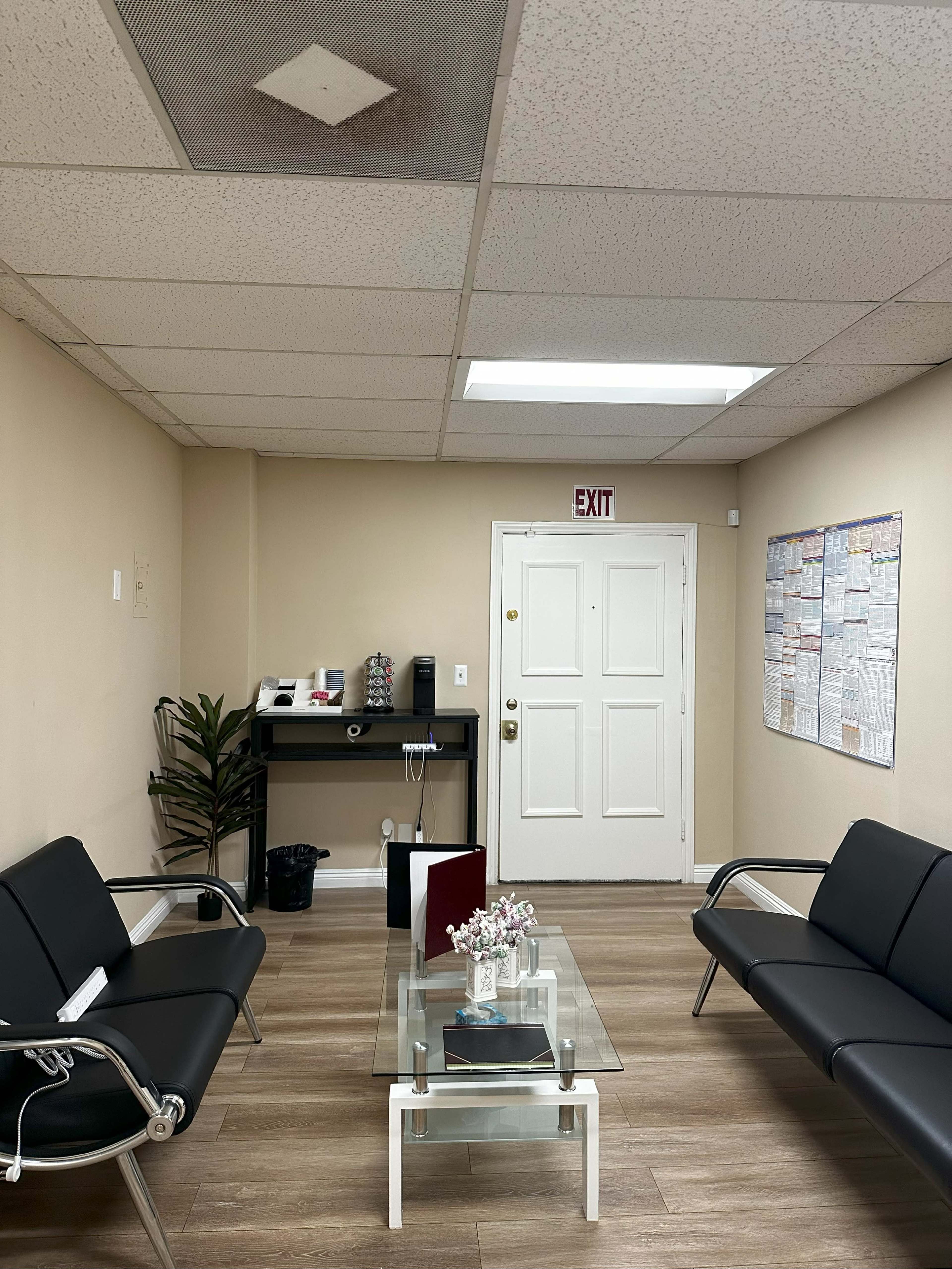 The image shows a waiting room with two black couches, a glass coffee table, a small potted plant, and a door leading to an exit.