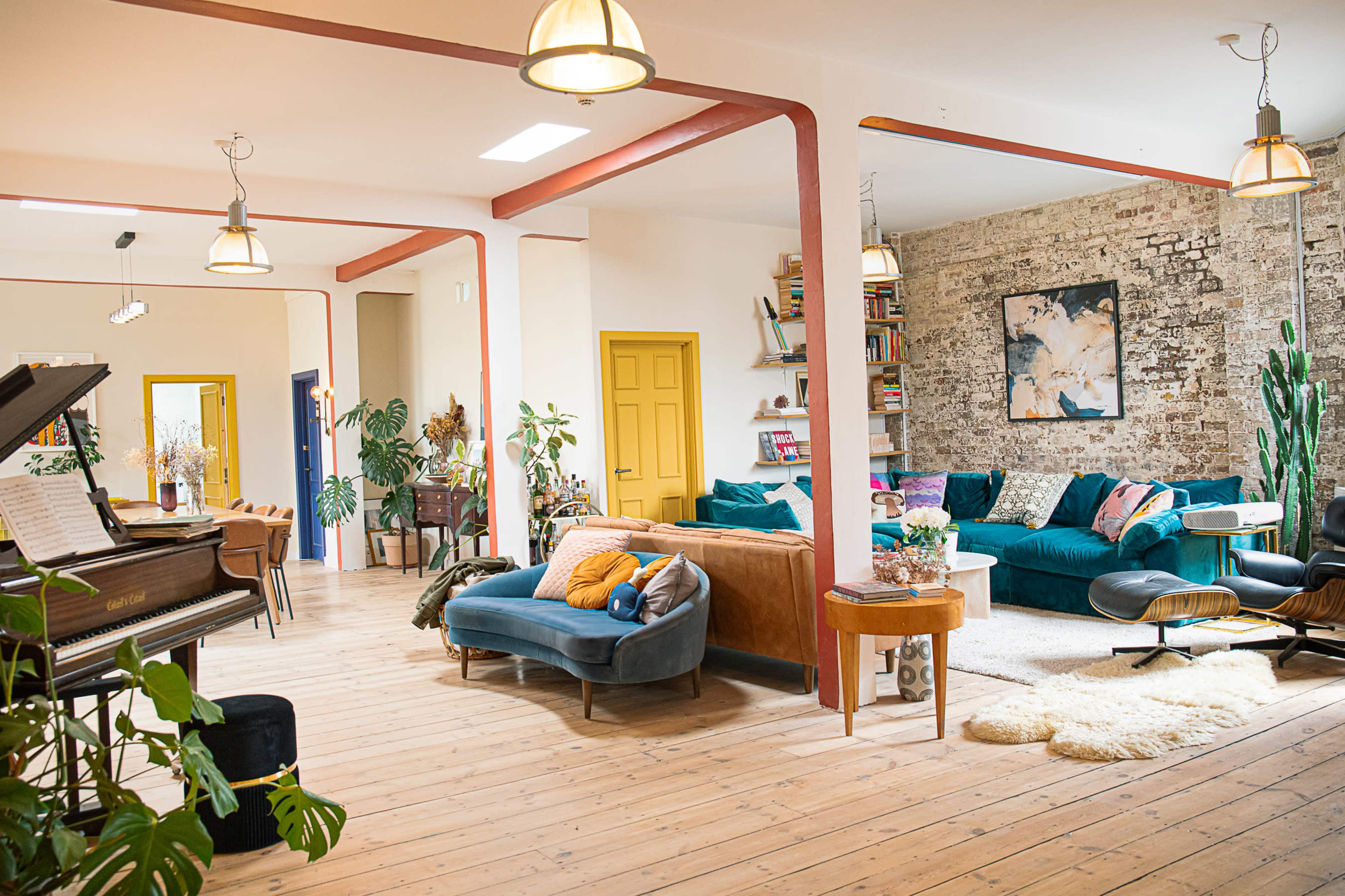 The image depicts a spacious, light-filled interior featuring a living area with multiple seating options, bookshelves, and a large piano.