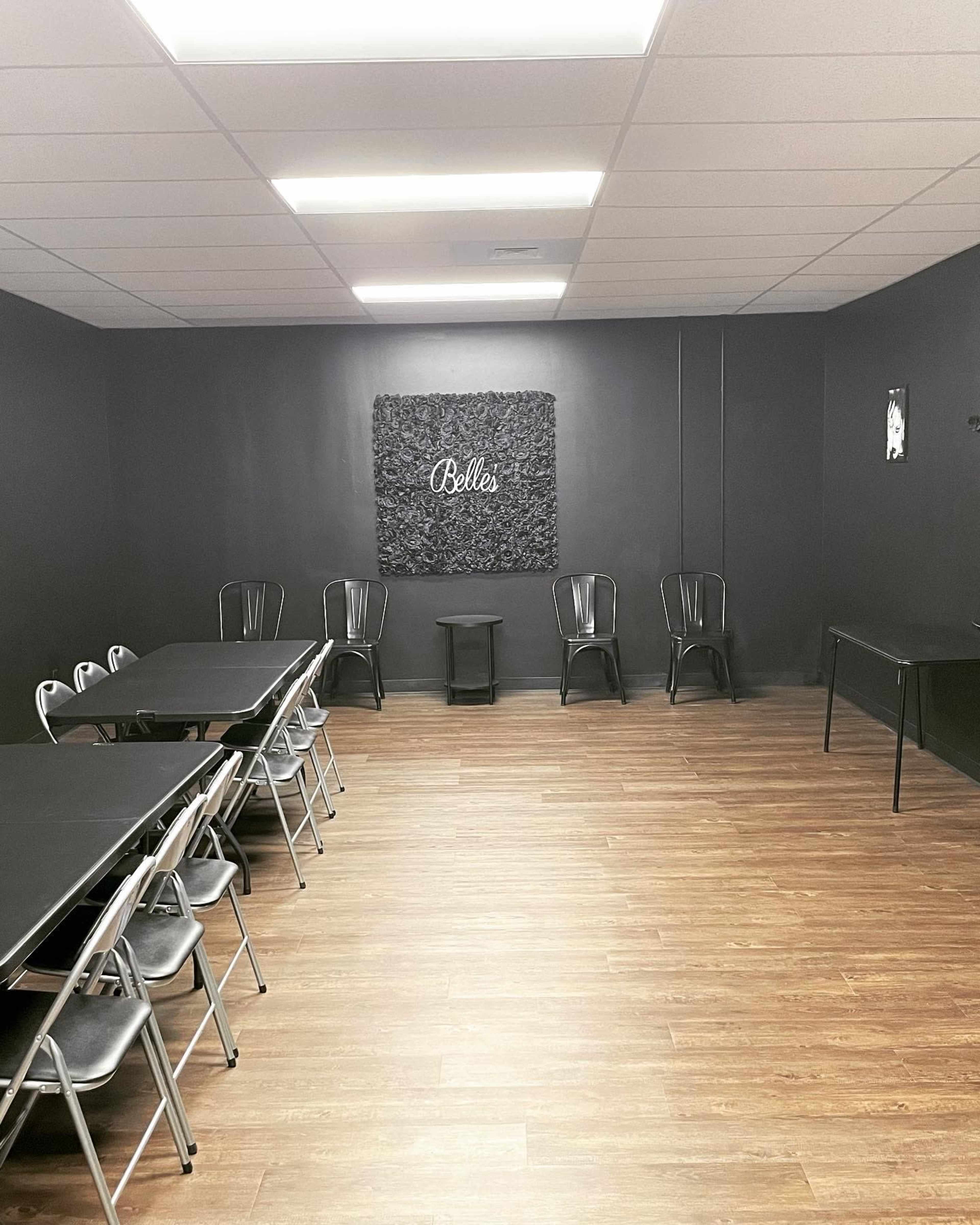The room features a dark wall with a decorative panel labeled "Belle's," two black tables, and several metal chairs arranged along the sides.