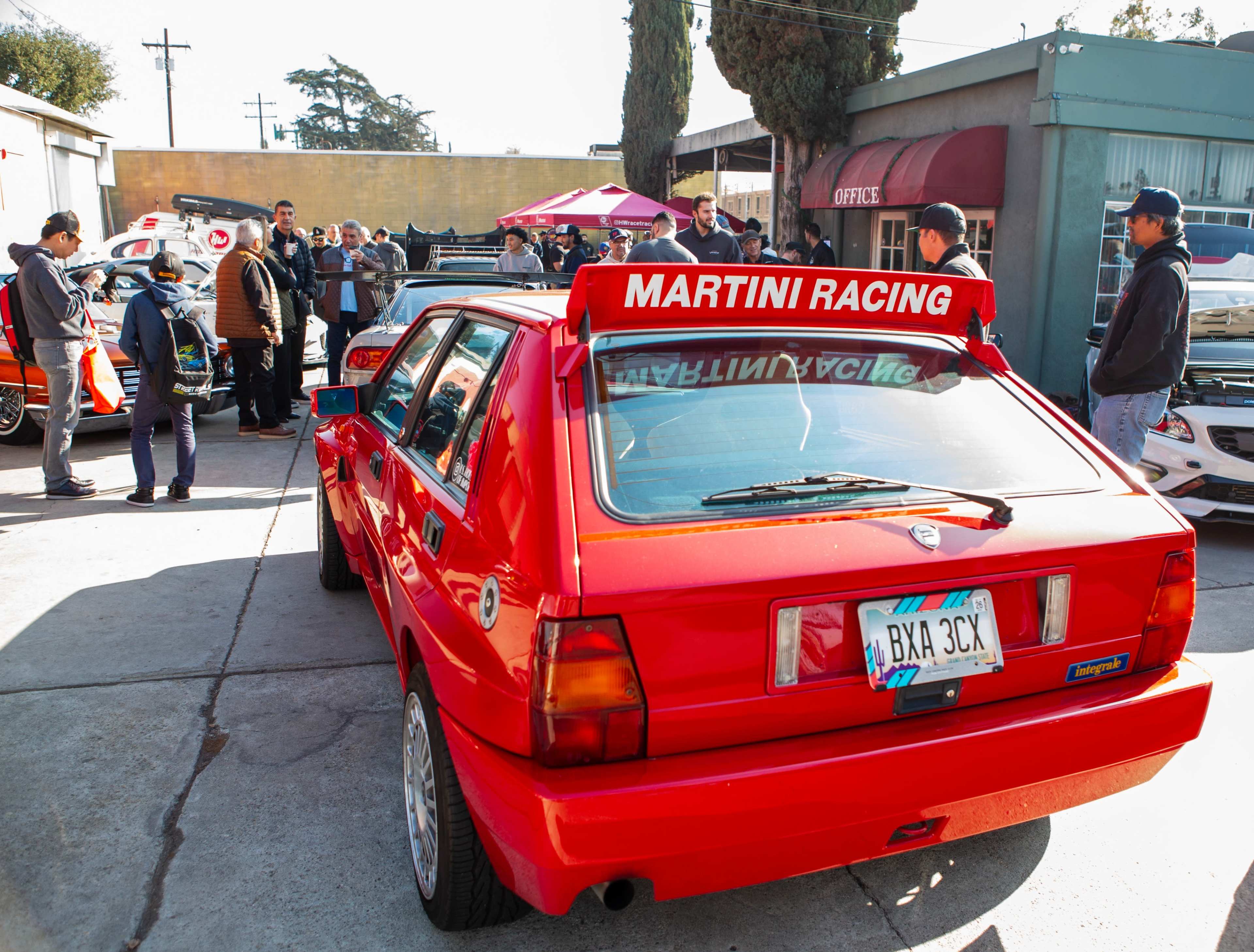 A red car with a "Martini Racing" sign sits in a parking lot surrounded by a gathering of people and several other vehicles.