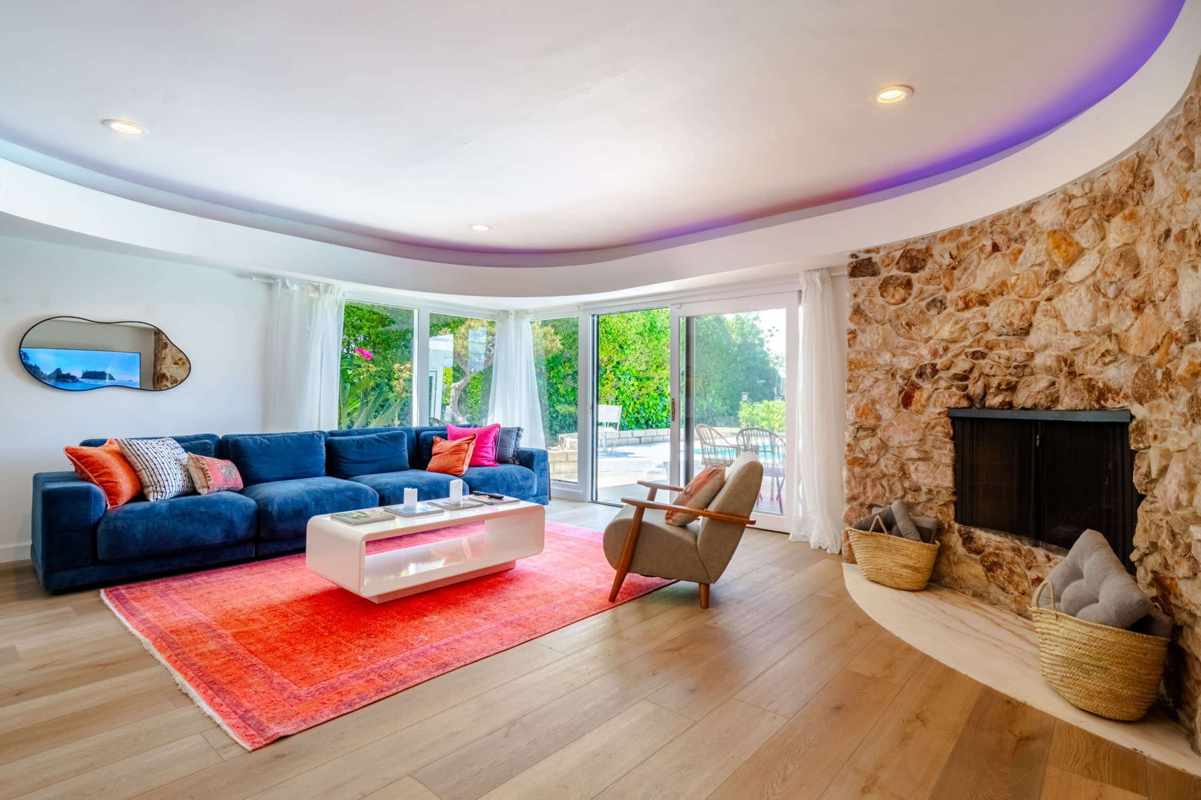 A modern living room featuring a blue sectional sofa, a white coffee table, a stone accent wall with a fireplace, and large windows that open to a garden view.