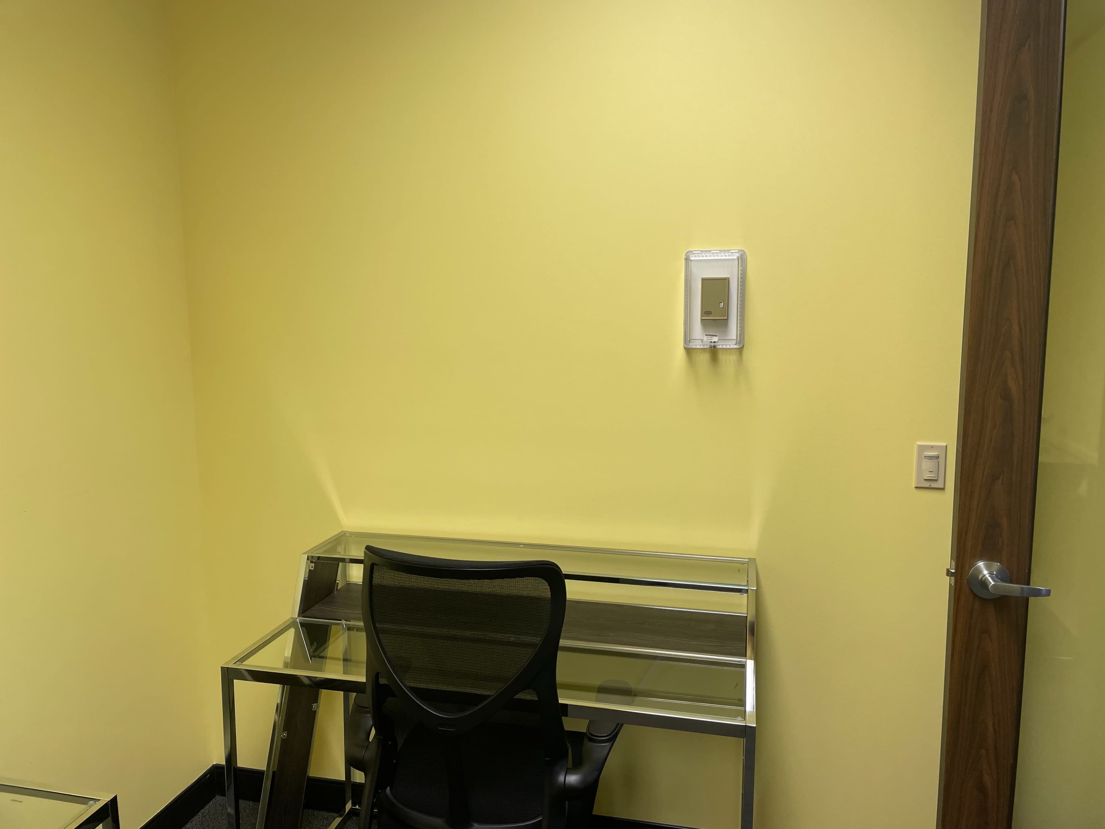 A small office space with a yellow wall, a glass desk, a black chair, and a door on the right.