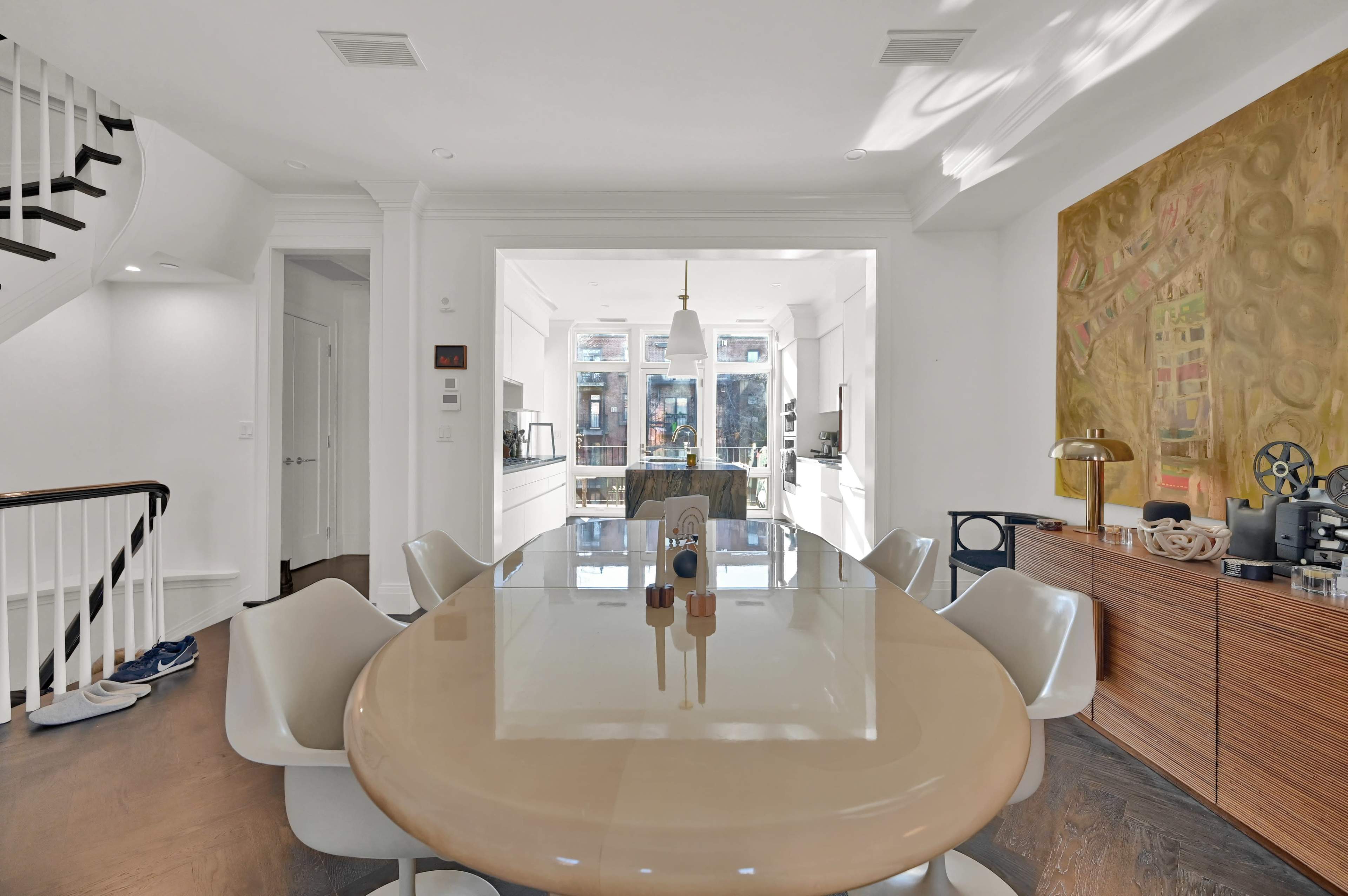A bright dining area features a large oval table surrounded by white chairs, with a wall art piece and a wooden sideboard in the background.