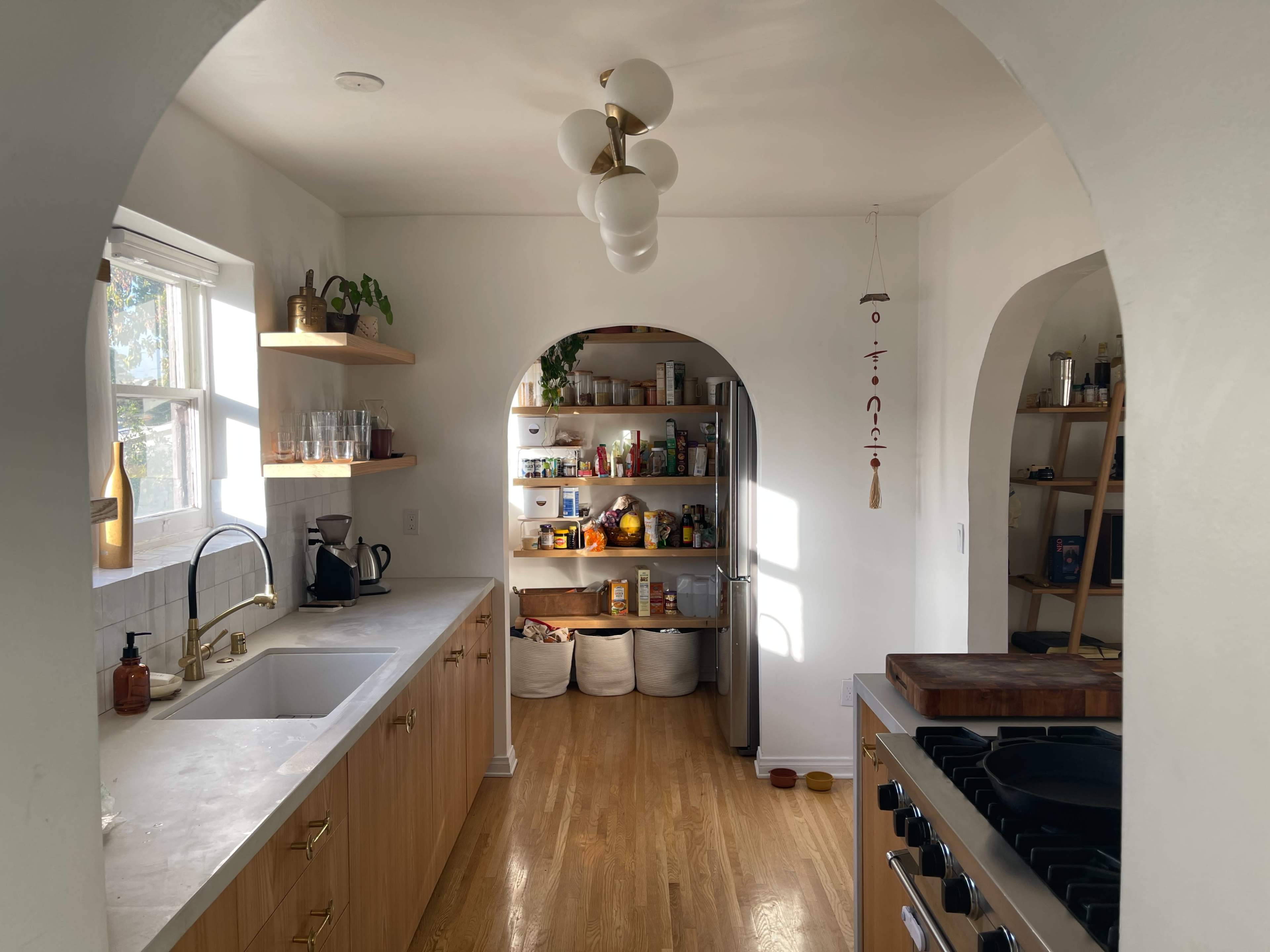 A bright kitchen features an archway leading to a pantry stocked with food items, with wood cabinets and a large sink along one side.