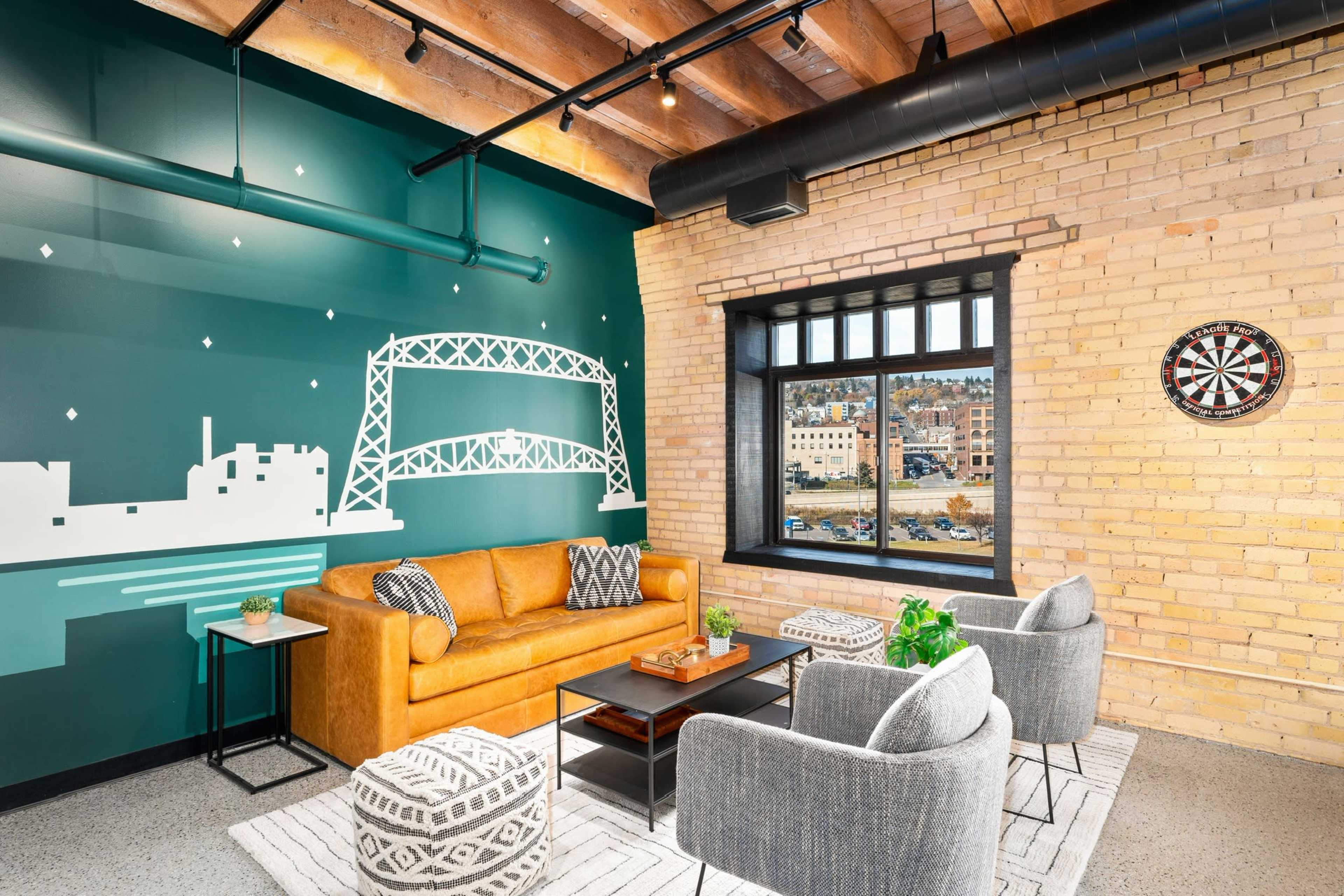 A cozy lounge area featuring a yellow sofa, two gray chairs, and a decorative wall with a mural of a bridge, all set against a backdrop of exposed brick and a window overlooking an outdoor scene.