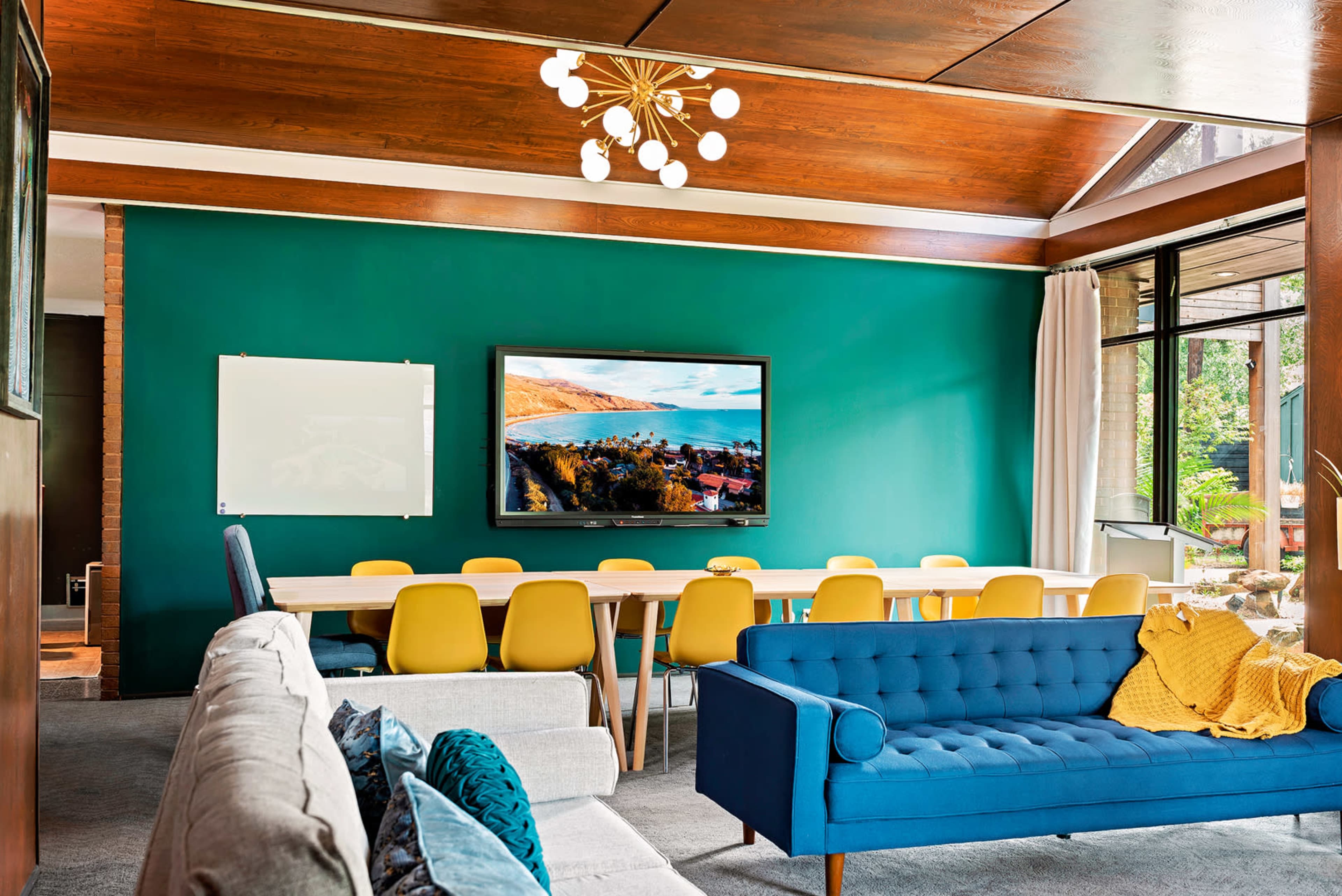 A spacious meeting room features a long table with yellow chairs, a large television on a green wall, and two sofas in the foreground.