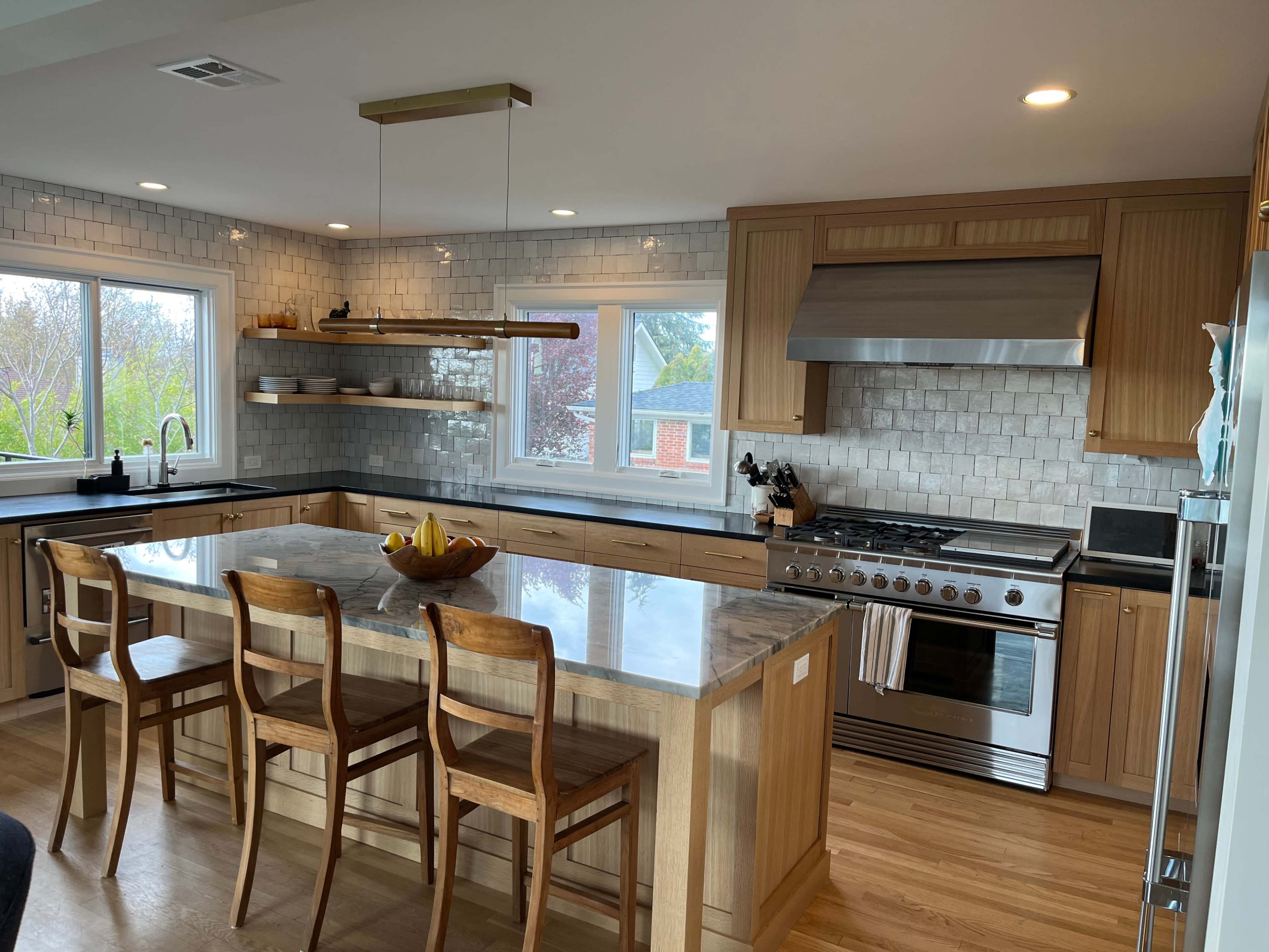 The image shows a modern kitchen featuring a large island with wooden bar stools, stainless steel appliances, and tiled backsplashes.