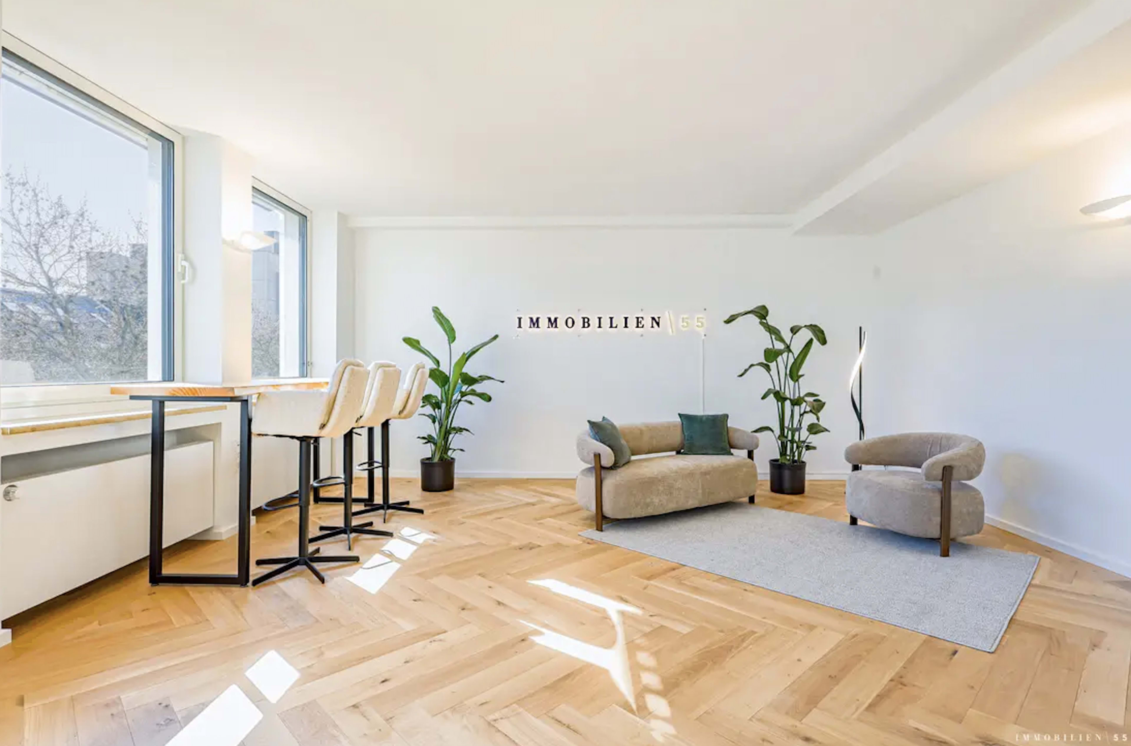 The image shows a modern, bright interior space with a seating area, a small rug, a table with high chairs, and several potted plants.