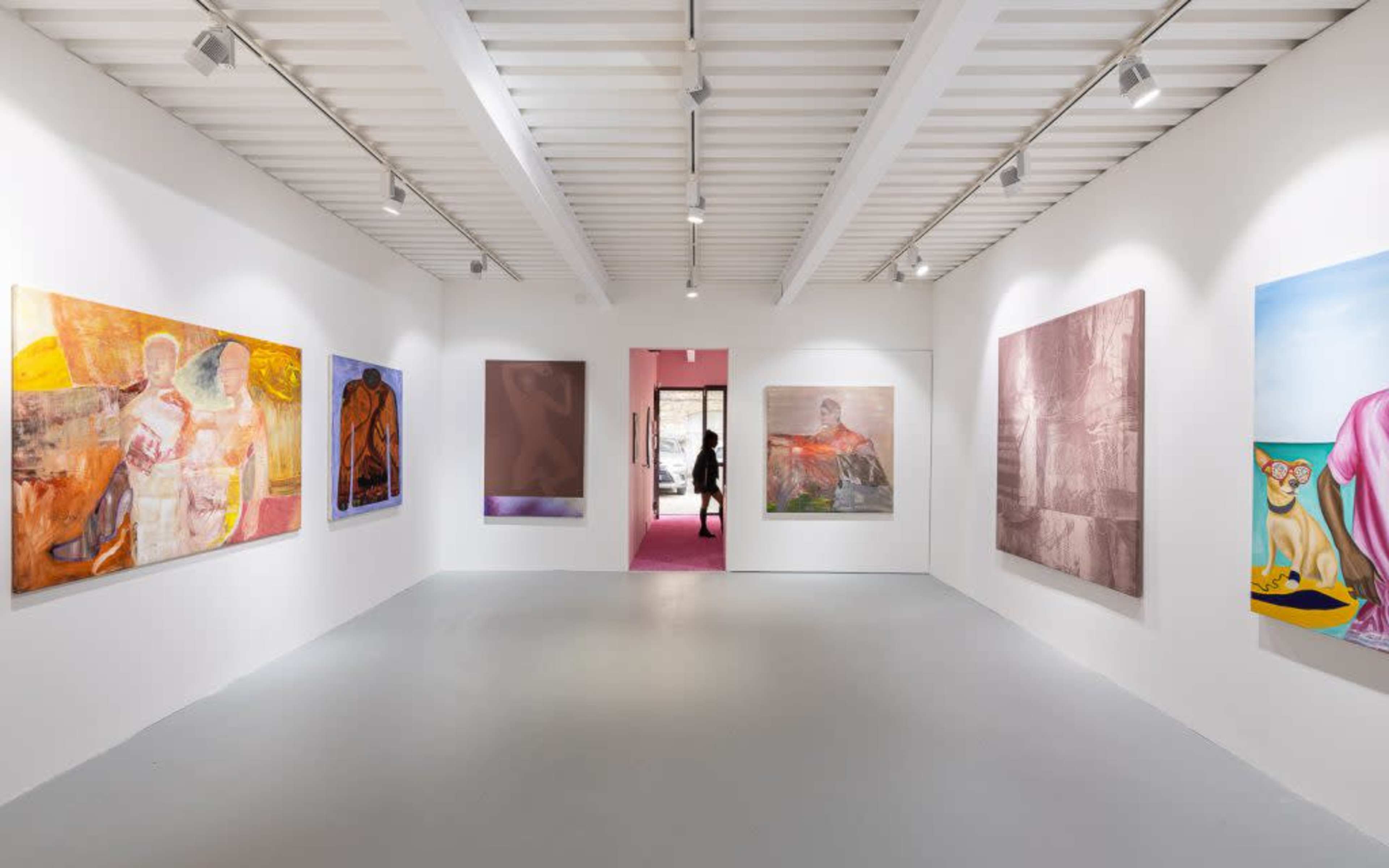 An art gallery with large, colorful paintings displayed on the white walls, and a person walking through a doorway at the far end.