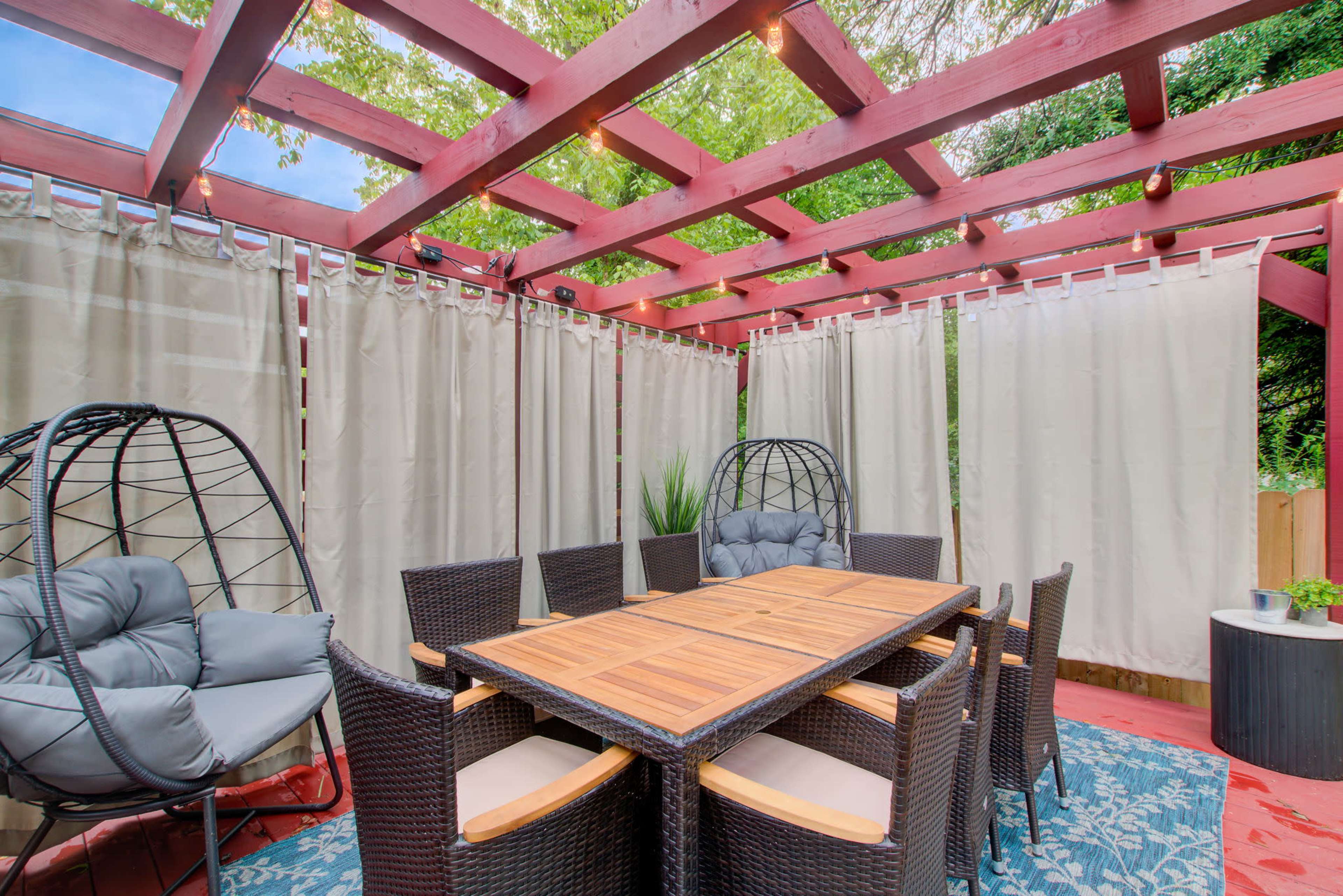 A covered outdoor dining area features a table surrounded by wicker chairs and two hanging chair swings, enclosed by sheer curtains.