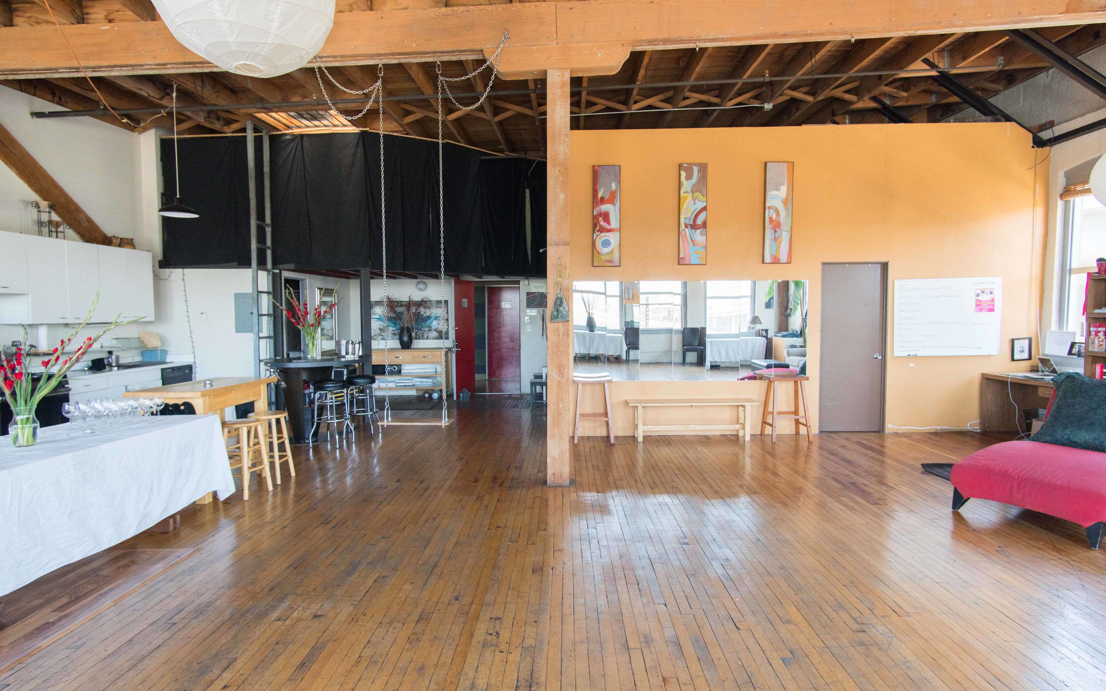 A spacious artist studio featuring wooden floors, exposed beams, a kitchenette area, and several pieces of abstract art on the walls.