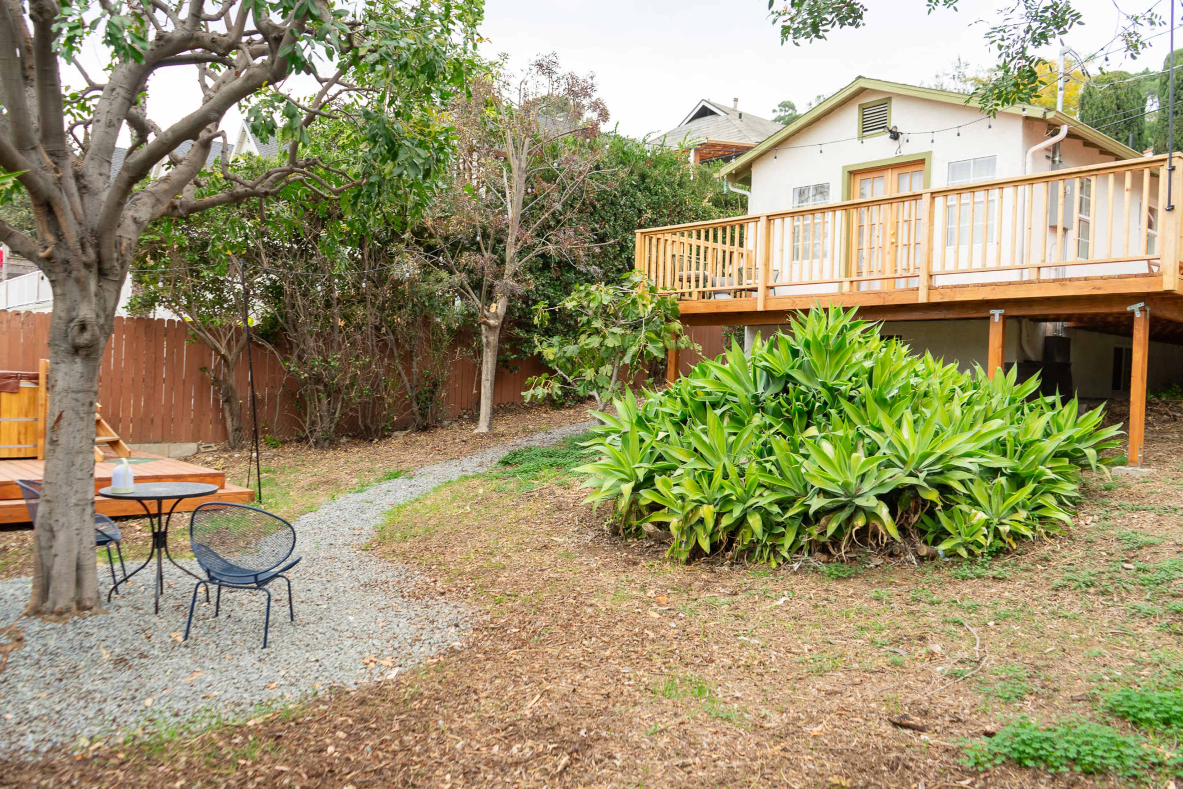 The image shows a backyard with a wooden deck attached to a house, surrounded by greenery and a gravel pathway leading to a seating area with a table and chair.