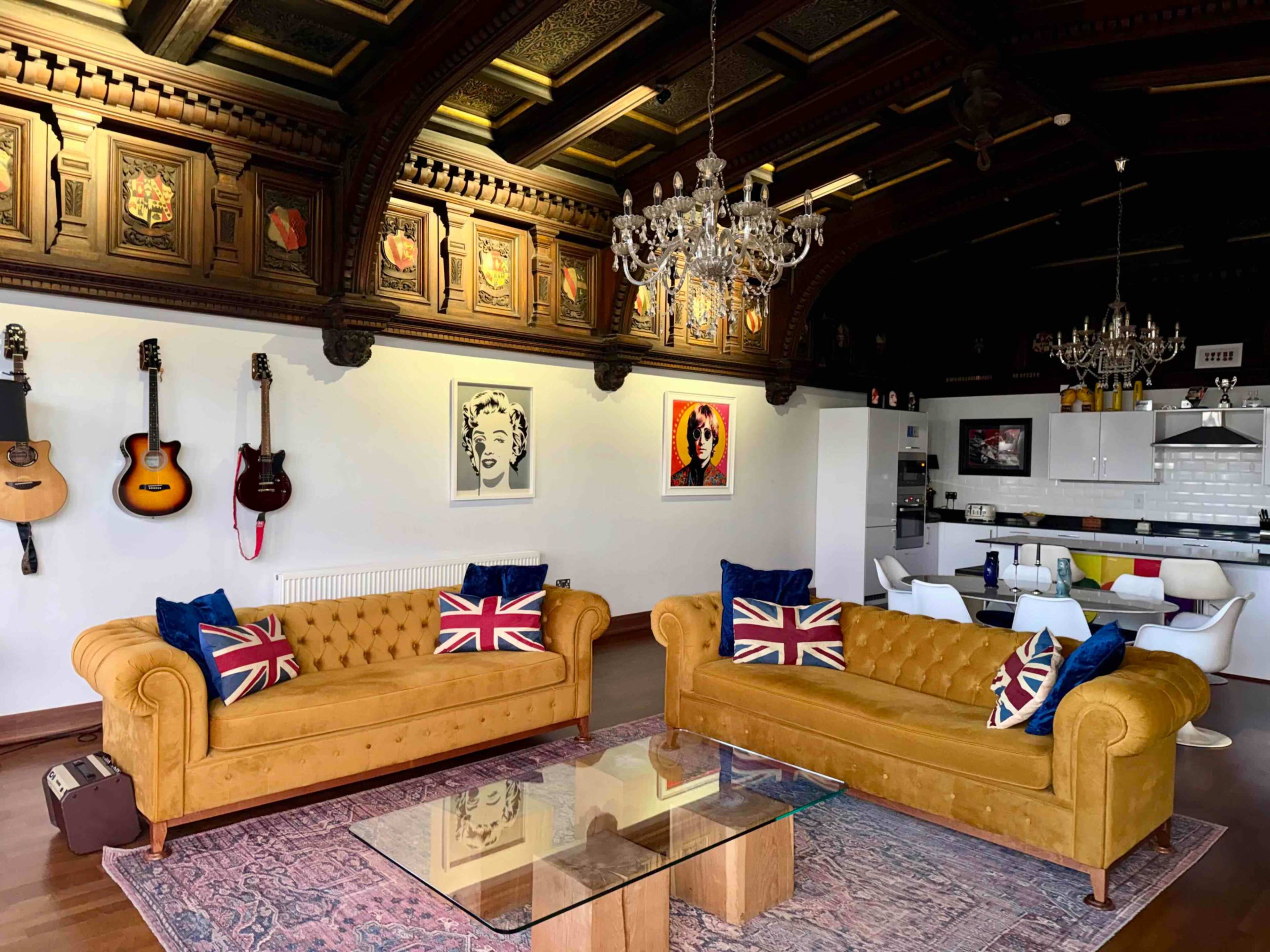 The image shows a stylish living area featuring two yellow Chesterfield sofas with Union Jack cushions, a glass coffee table, and a backdrop of wooden paneling, guitars, and framed artworks.
