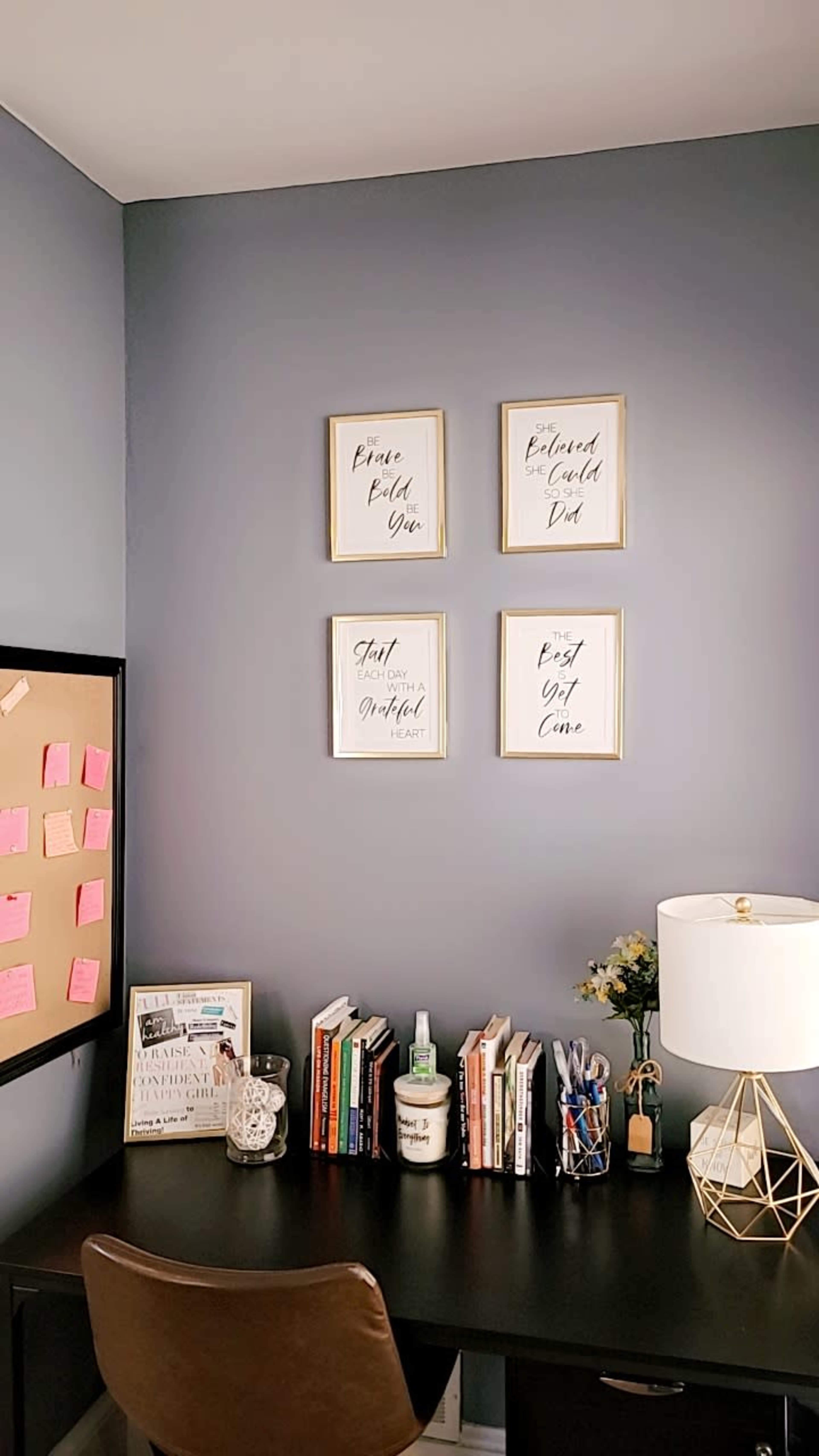 The image shows a home office corner with a dark desk, a wall adorned with framed quotes, a bulletin board covered in notes, and a small bookshelf filled with books.