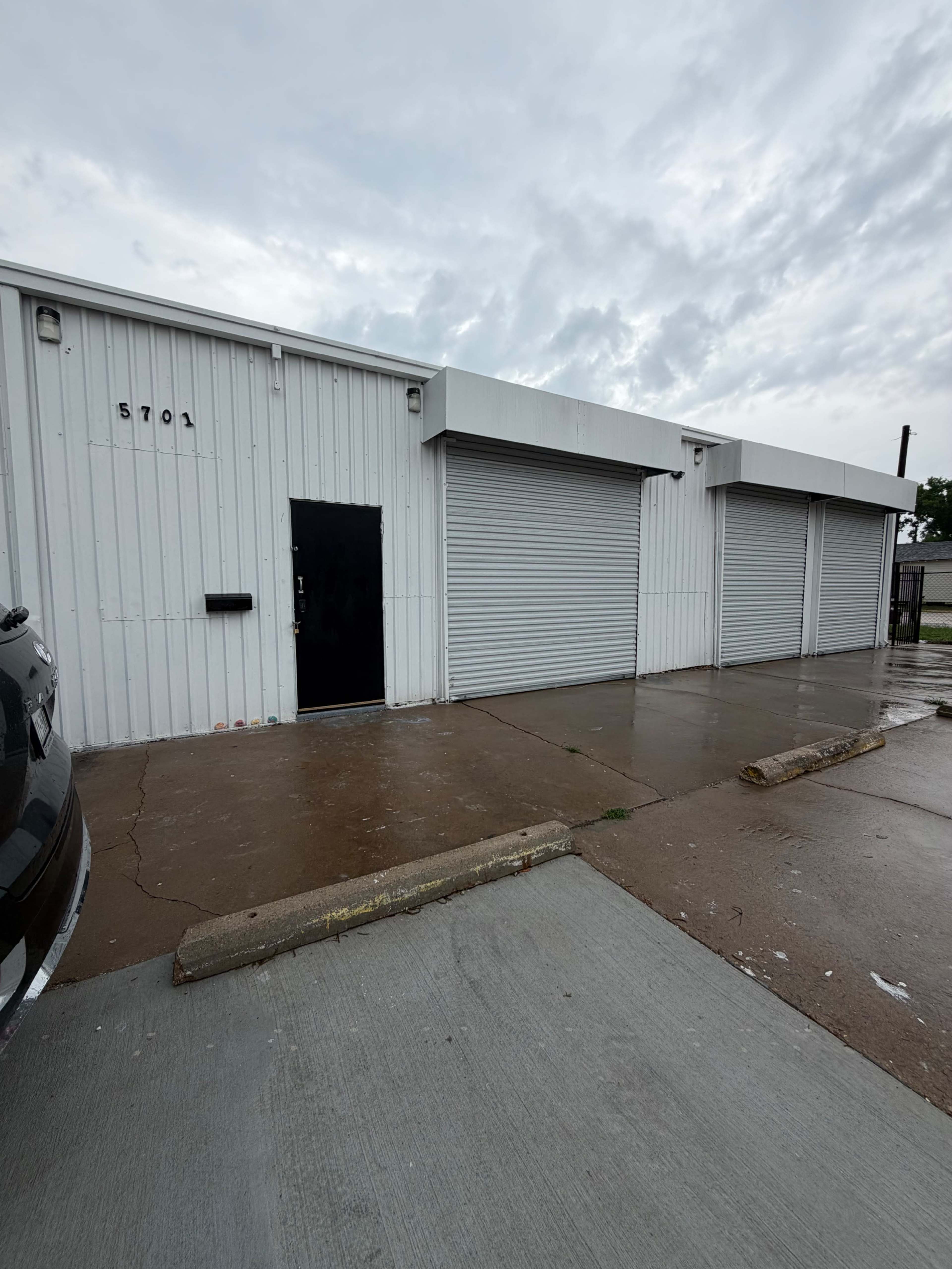 The image shows a single-story industrial building with a metal exterior, featuring a door and three roll-up garage doors, set on a wet concrete surface.