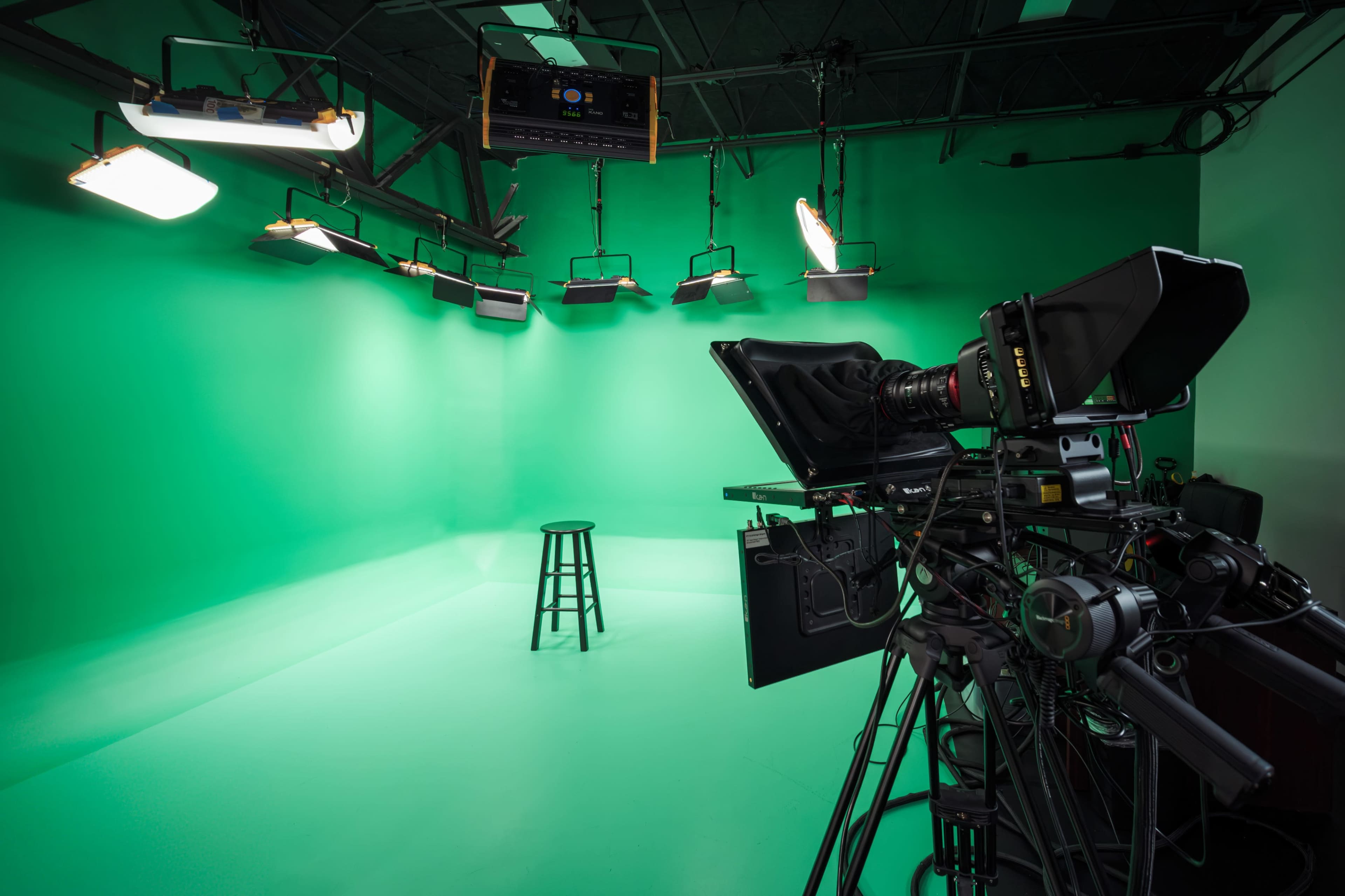 A video production studio features a green screen backdrop, a lighting setup, and a camera positioned beside an empty stool.