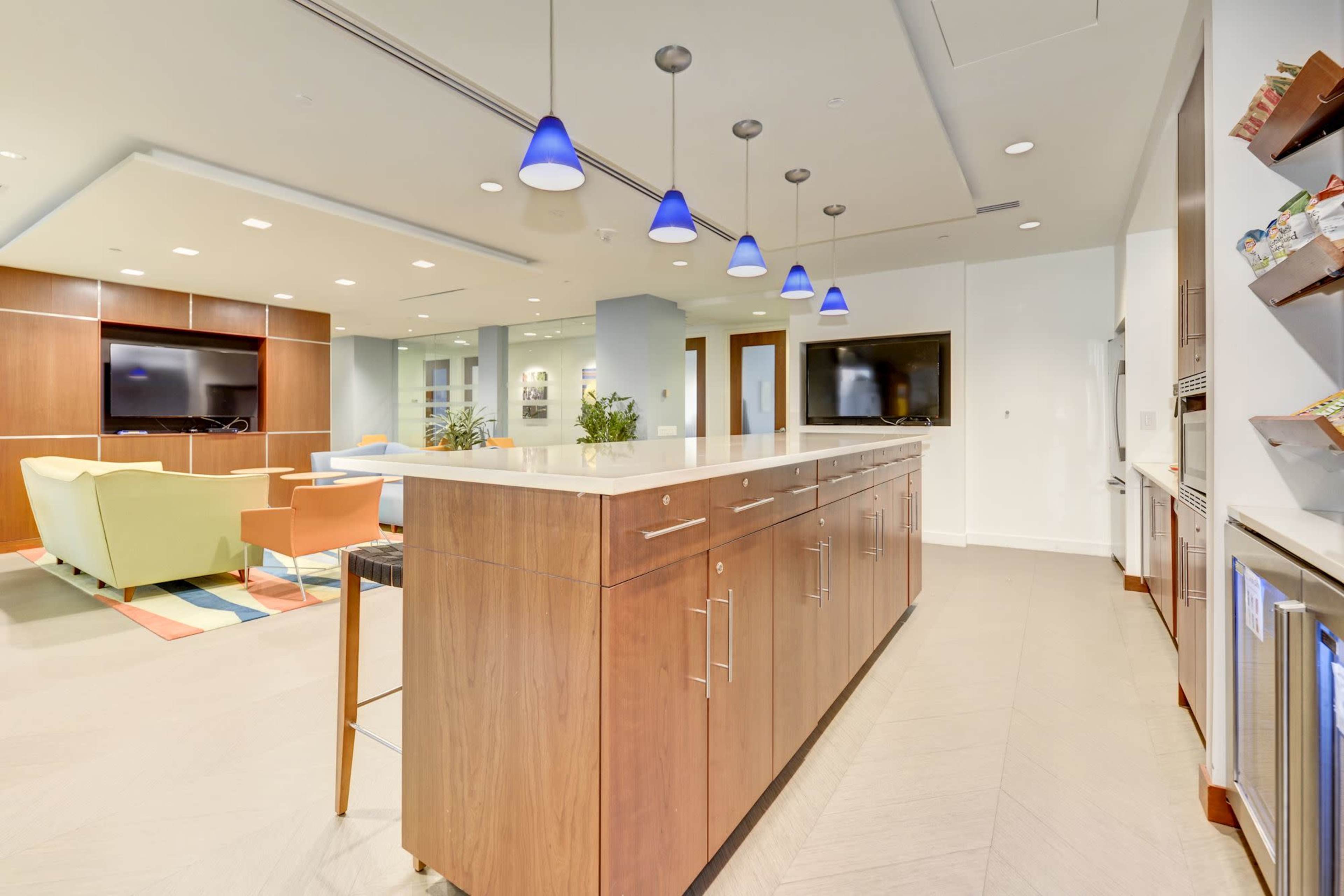 The image shows a modern kitchen-like space with a long wooden counter, pendant lighting, and various seating areas in a well-lit communal area.