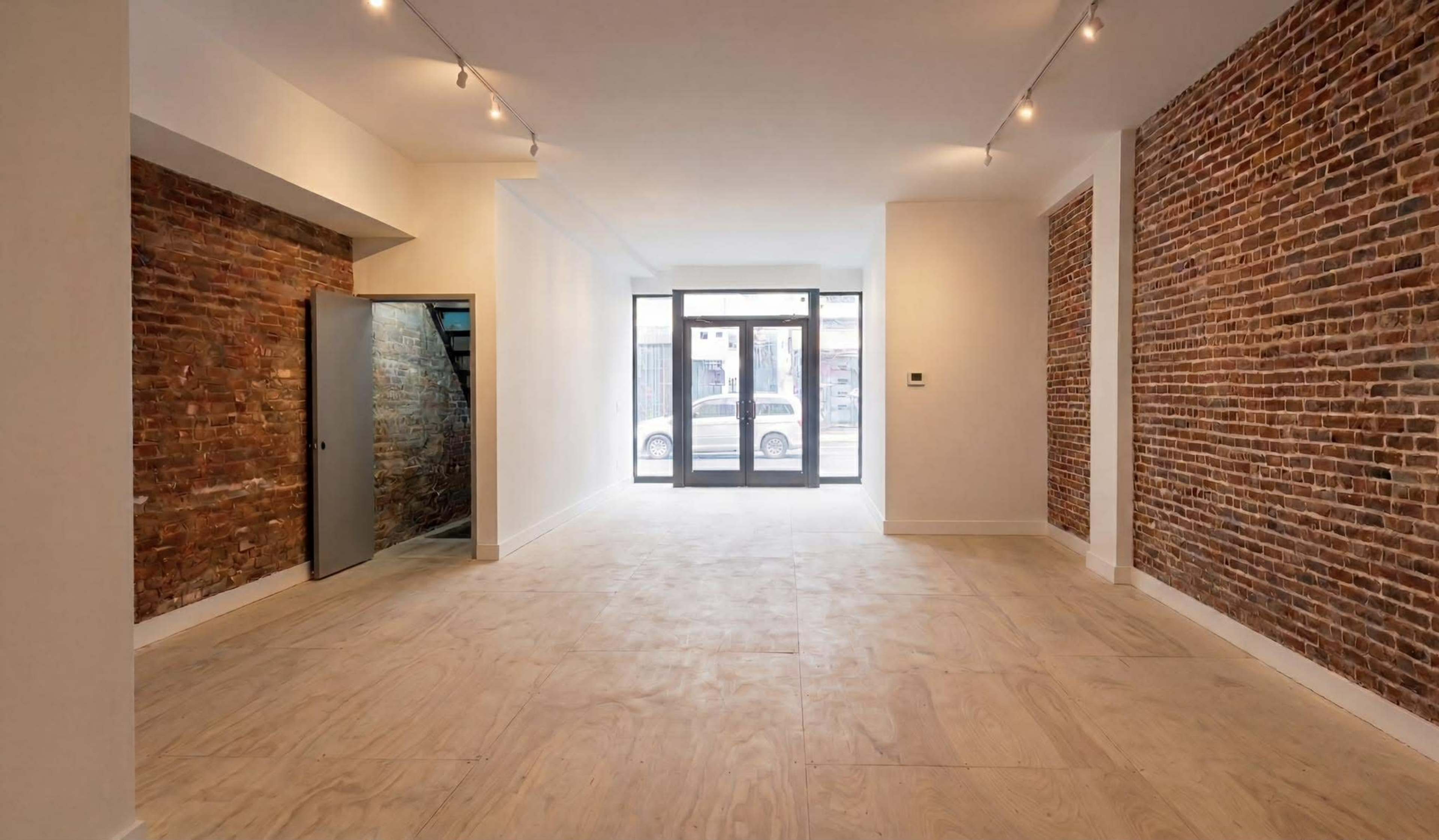 The image shows an empty interior space with exposed brick walls, wood flooring, and large windows facing the street.