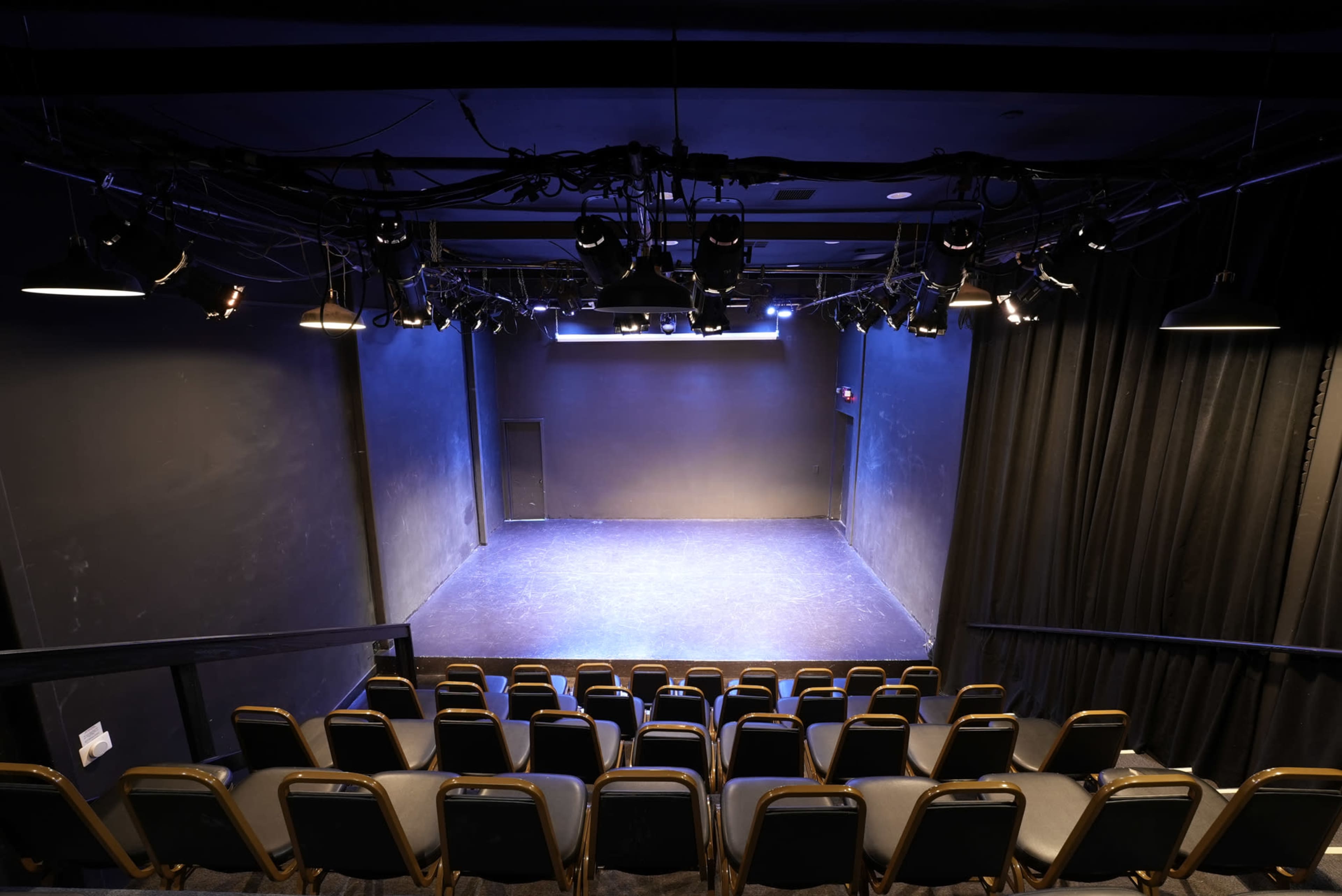 A small theater stage is illuminated by ceiling lights, with empty rows of seats facing the performance area.