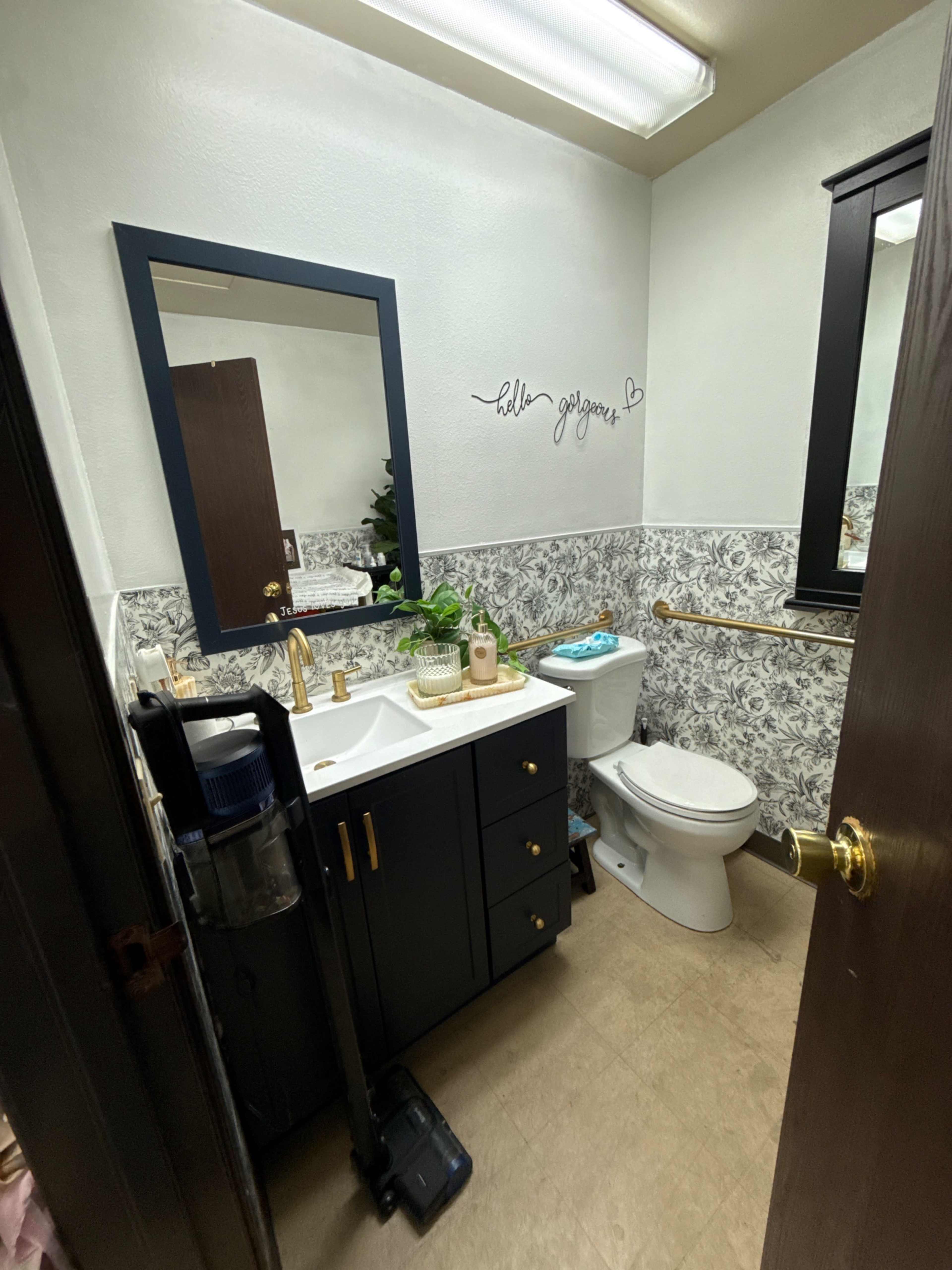 The image shows a small bathroom featuring a vanity with a sink, a toilet, a dark-colored mirror, and floral wallpaper on one wall.