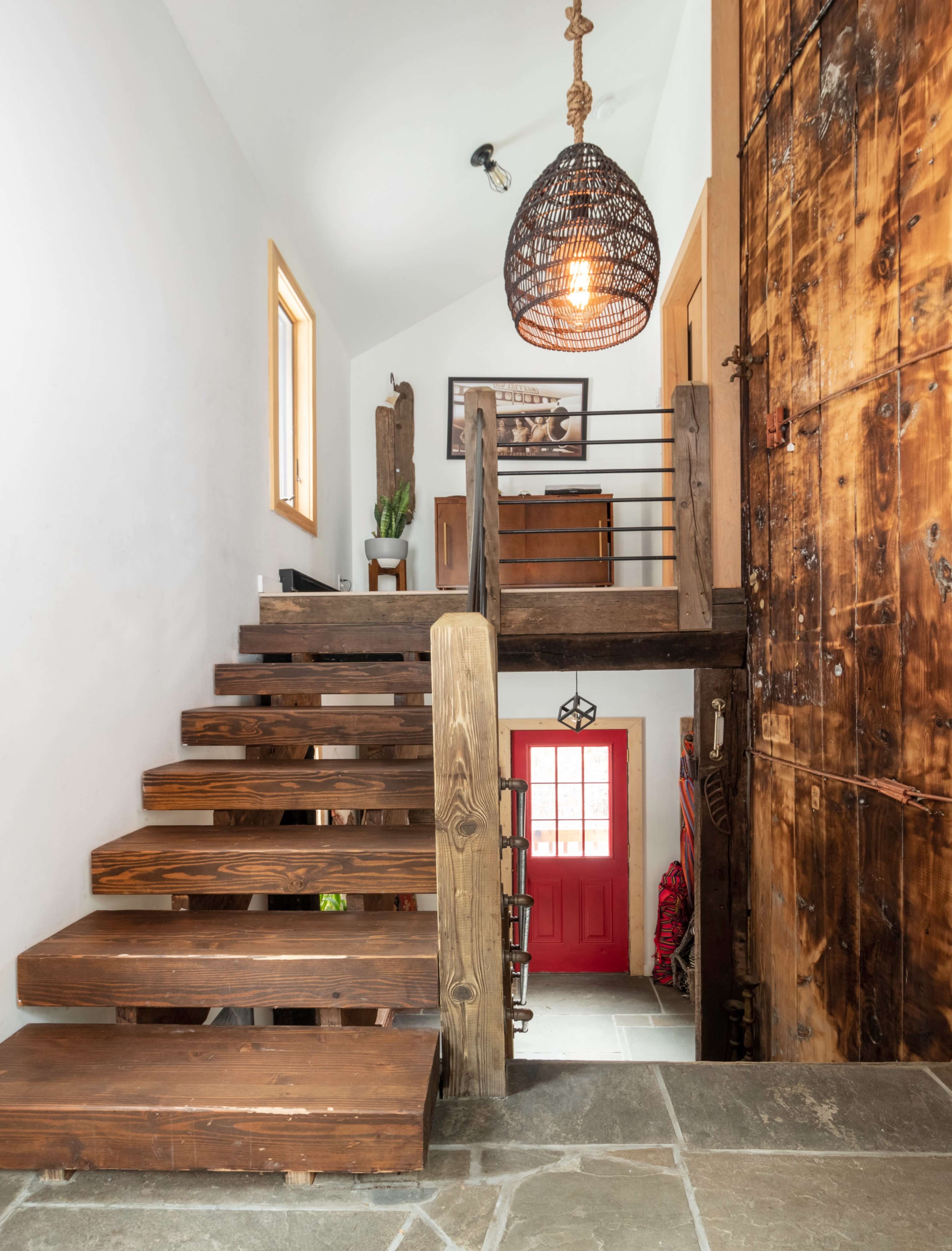 The image shows a wooden staircase leading to an upper level, with a large wooden door and a bright red entrance door visible at the bottom.