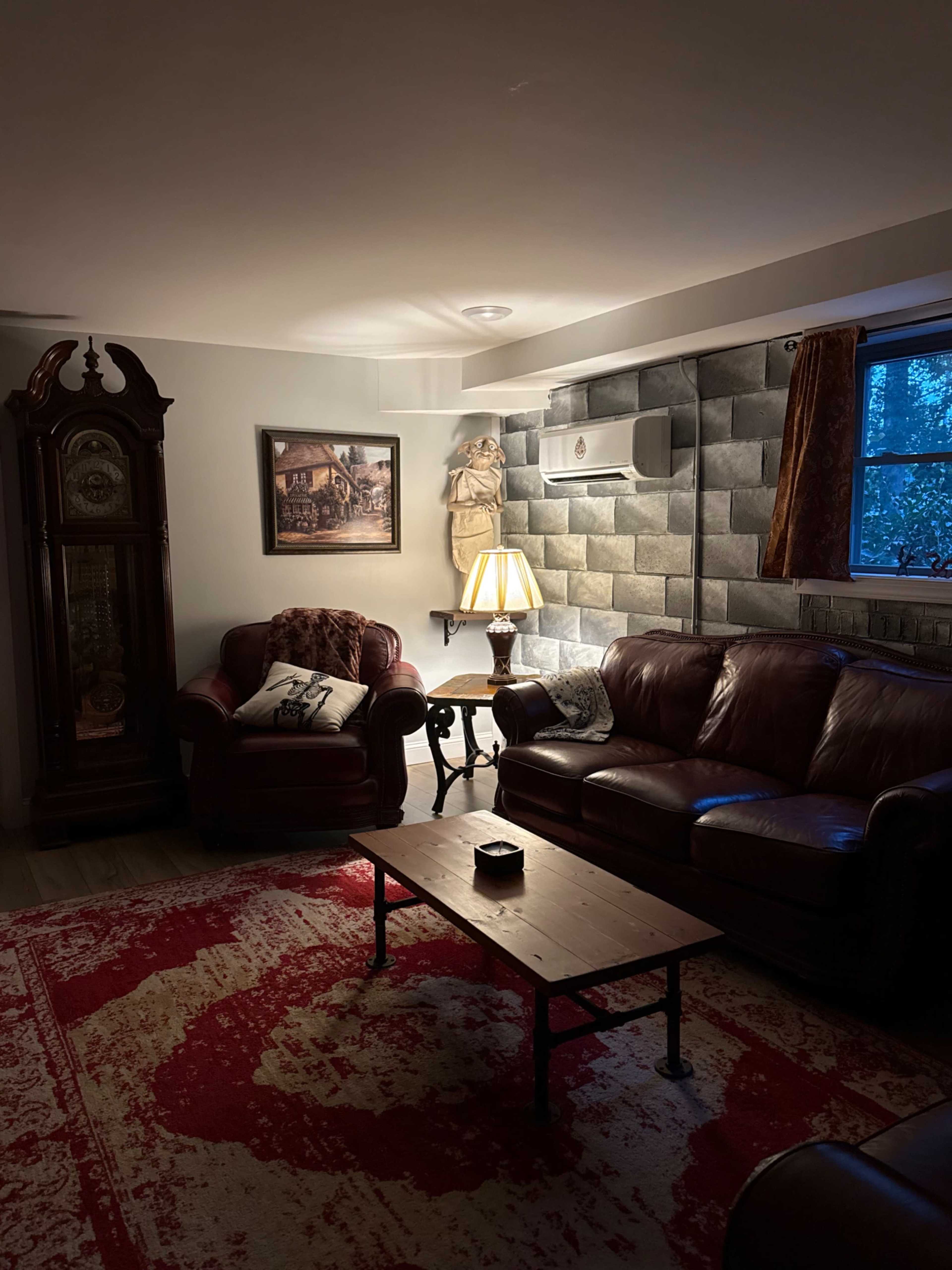 The room features leather couches, a wooden coffee table, a tall grandfather clock, and a decorative lamp, all situated against a stone wall.