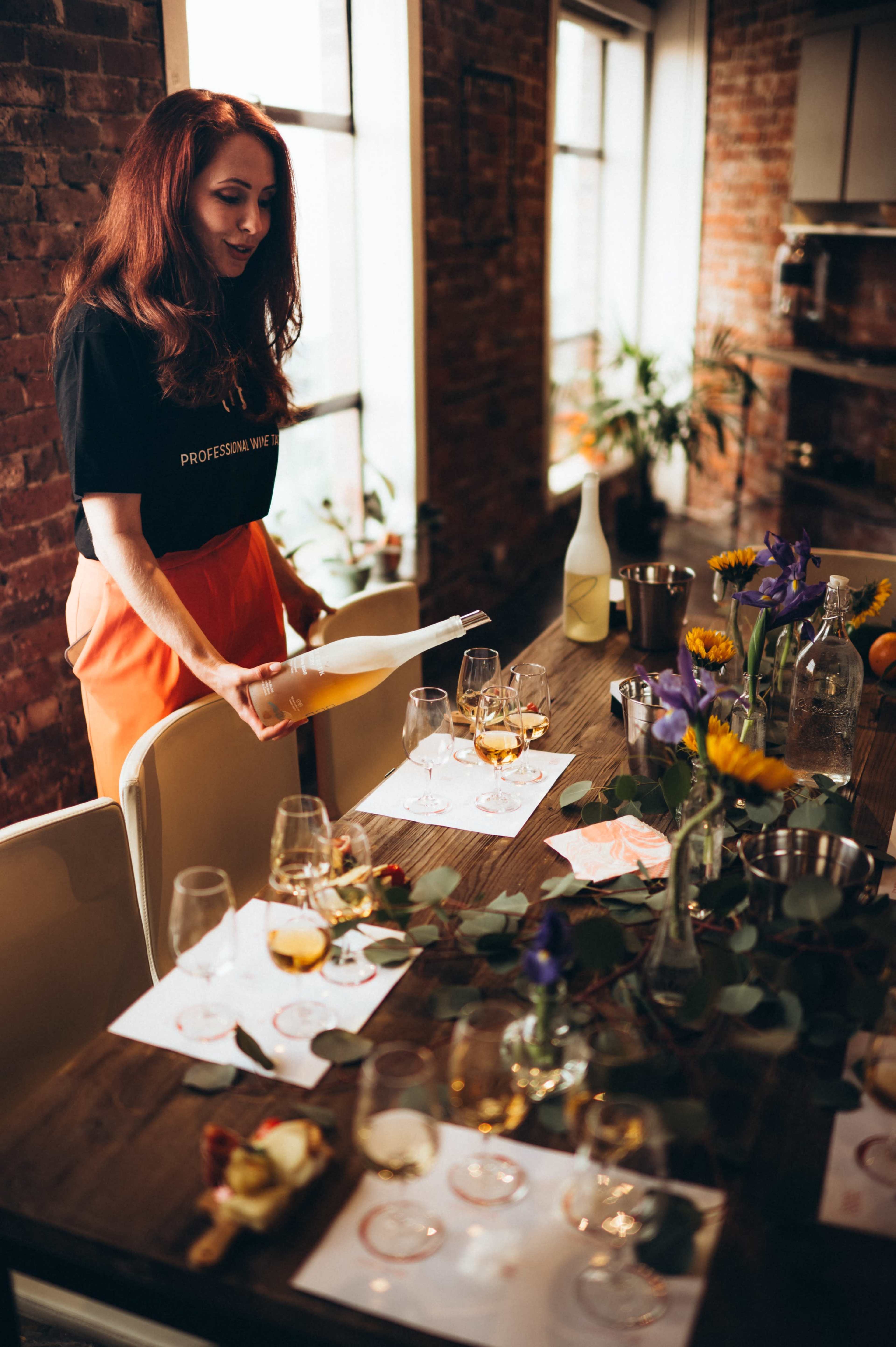 A woman in a black shirt prepares a table set with glasses and flowers in a brick-walled interior.