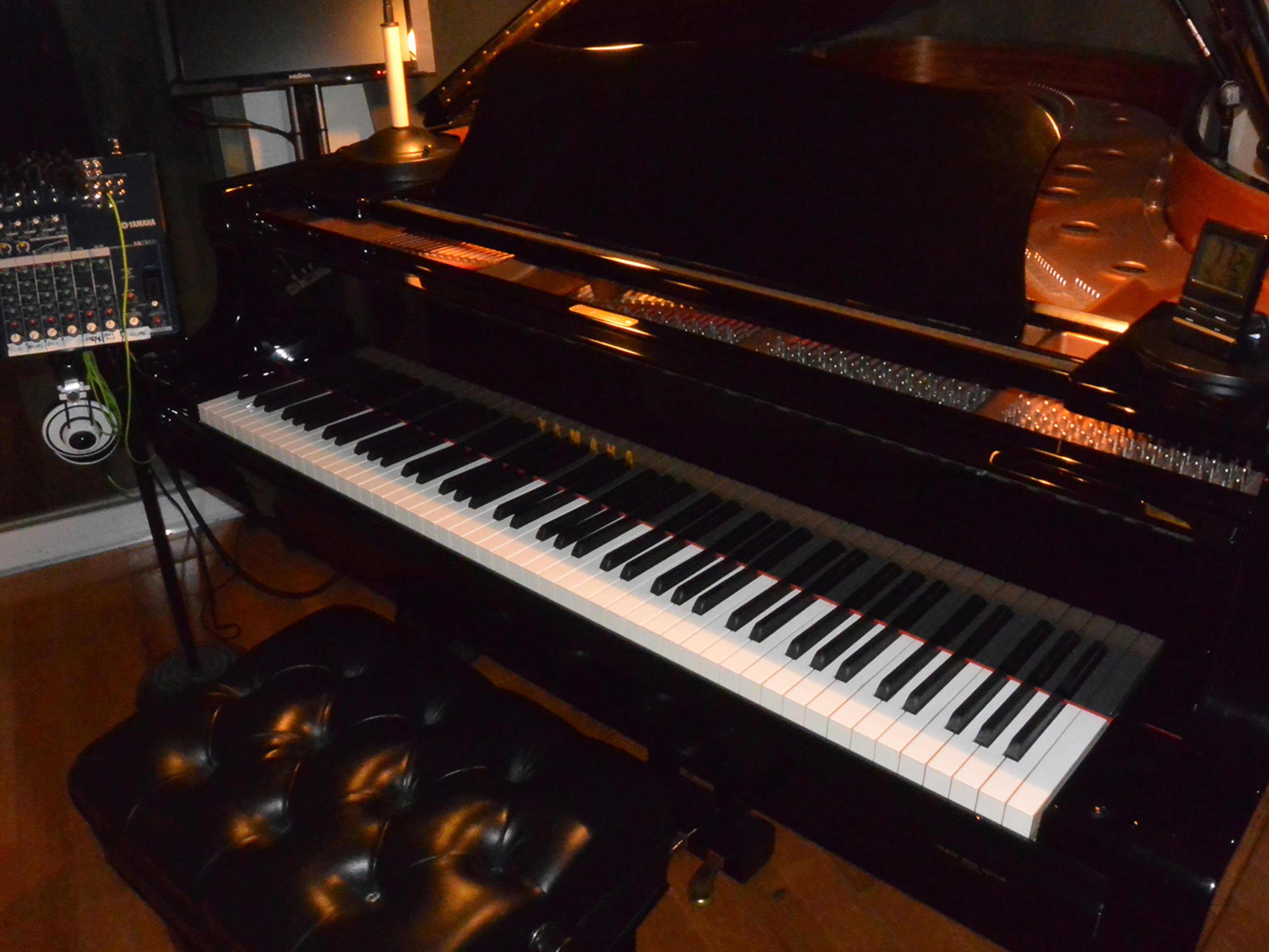 A black grand piano is positioned in a dimly lit room next to a sound mixing console and a black cushioned piano bench.