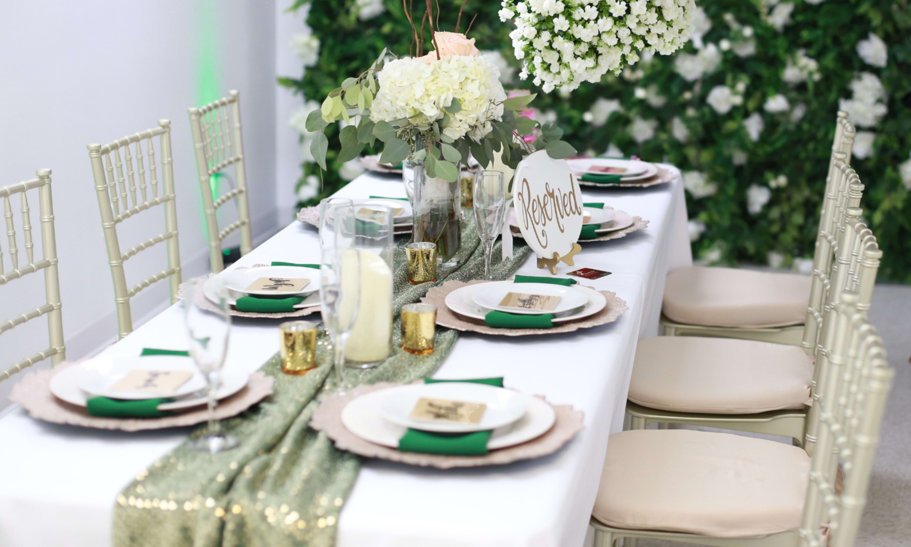 A elegantly set dining table features white tablecloths, gold and green accents, floral centerpieces, and chiavari chairs against a backdrop of greenery and white flowers.