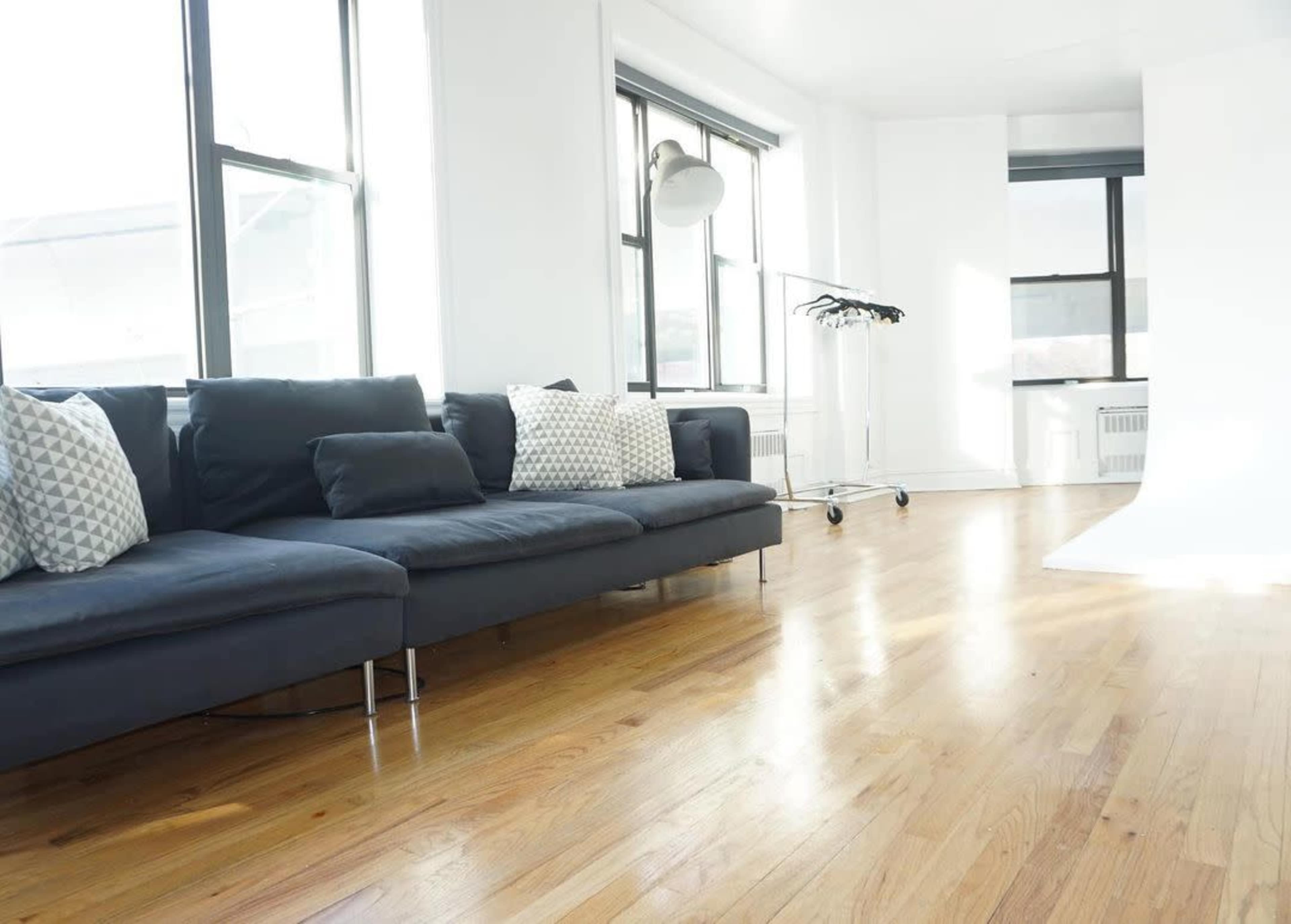 A bright, modern living space with a dark gray sectional sofa, decorative pillows, and a clothing rack beside large windows.
