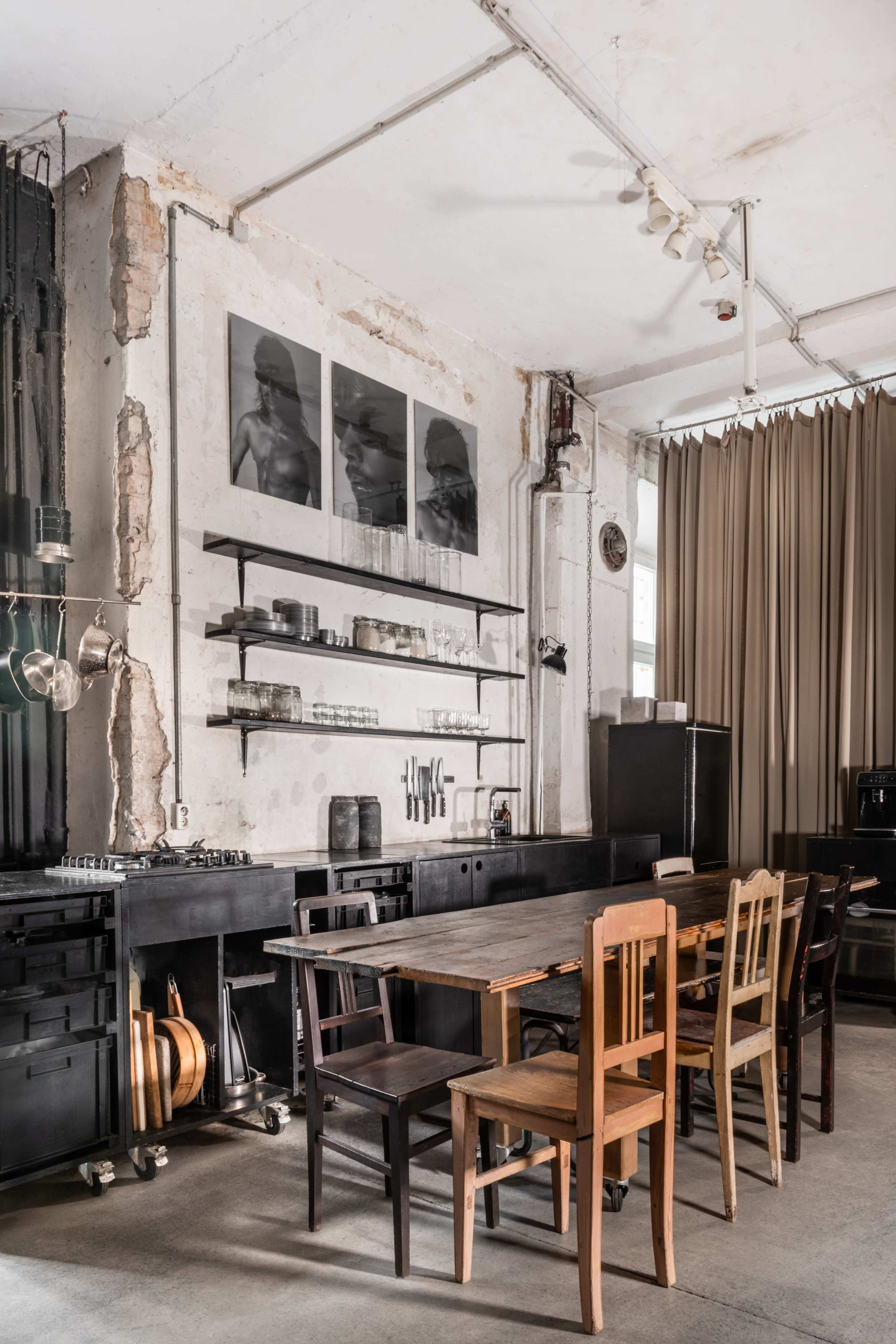 A minimalist kitchen with a long black table, various wooden chairs, and wall-mounted shelves displaying kitchenware and artwork.