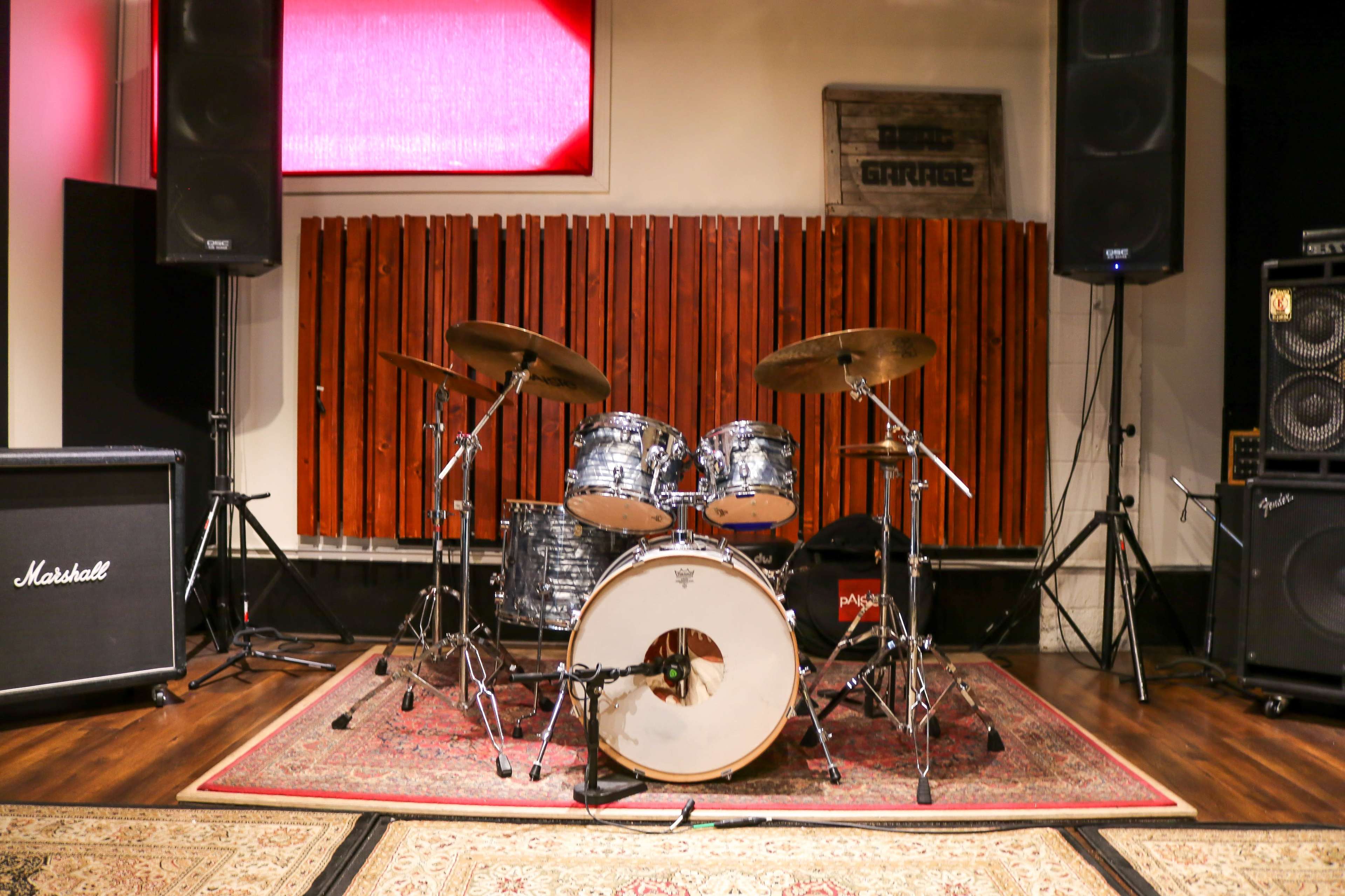A drum set is positioned in front of sound equipment on a wooden floor, with soundproofing panels on the wall behind it.
