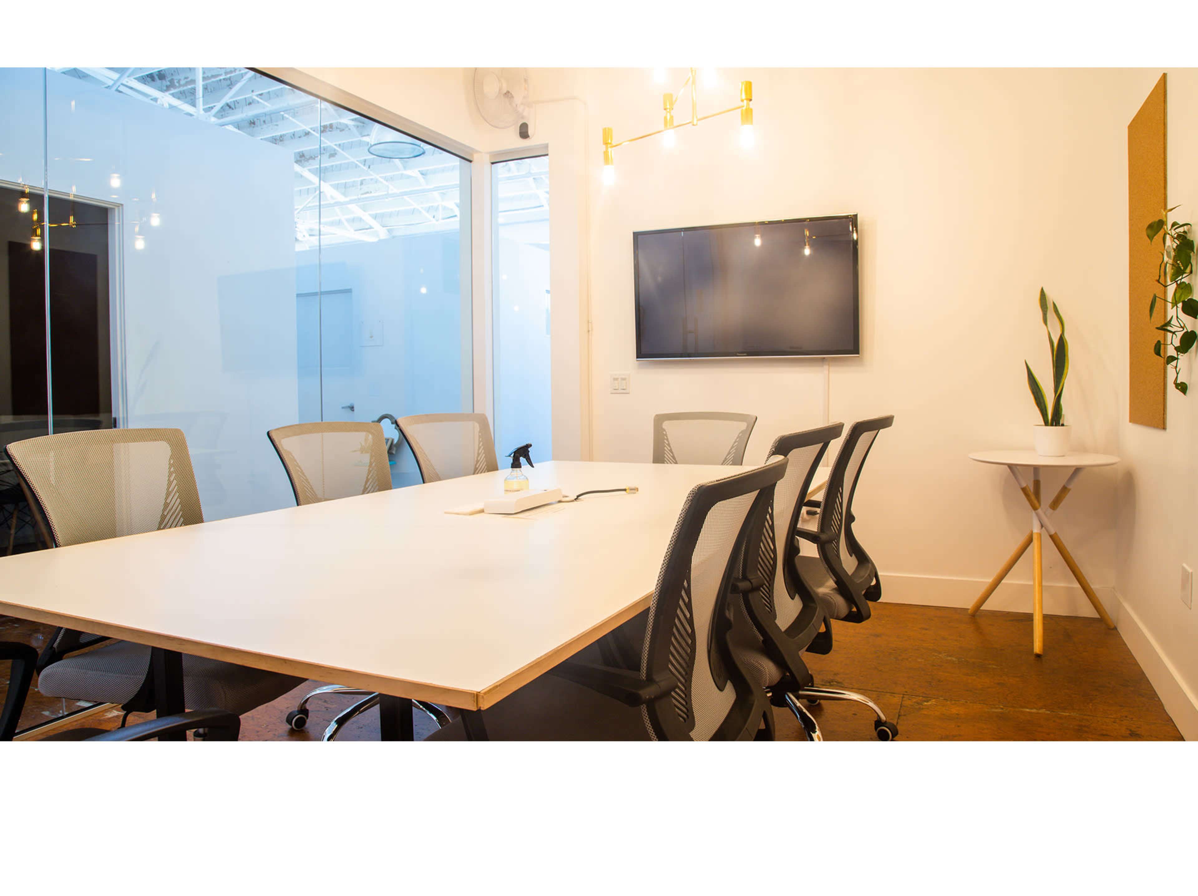 A modern conference room features a long white table surrounded by ergonomic chairs, a wall-mounted television, and a small plant on a side table.