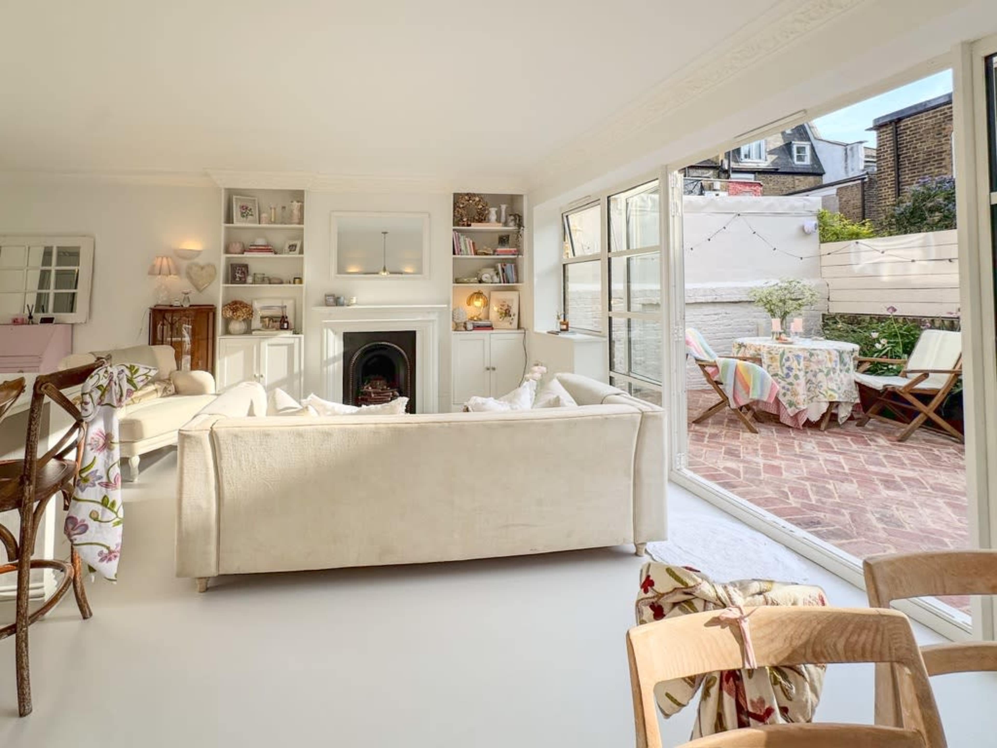 A bright living room features a white sofa, a fireplace, and large glass doors leading to a patio with outdoor seating and a table.