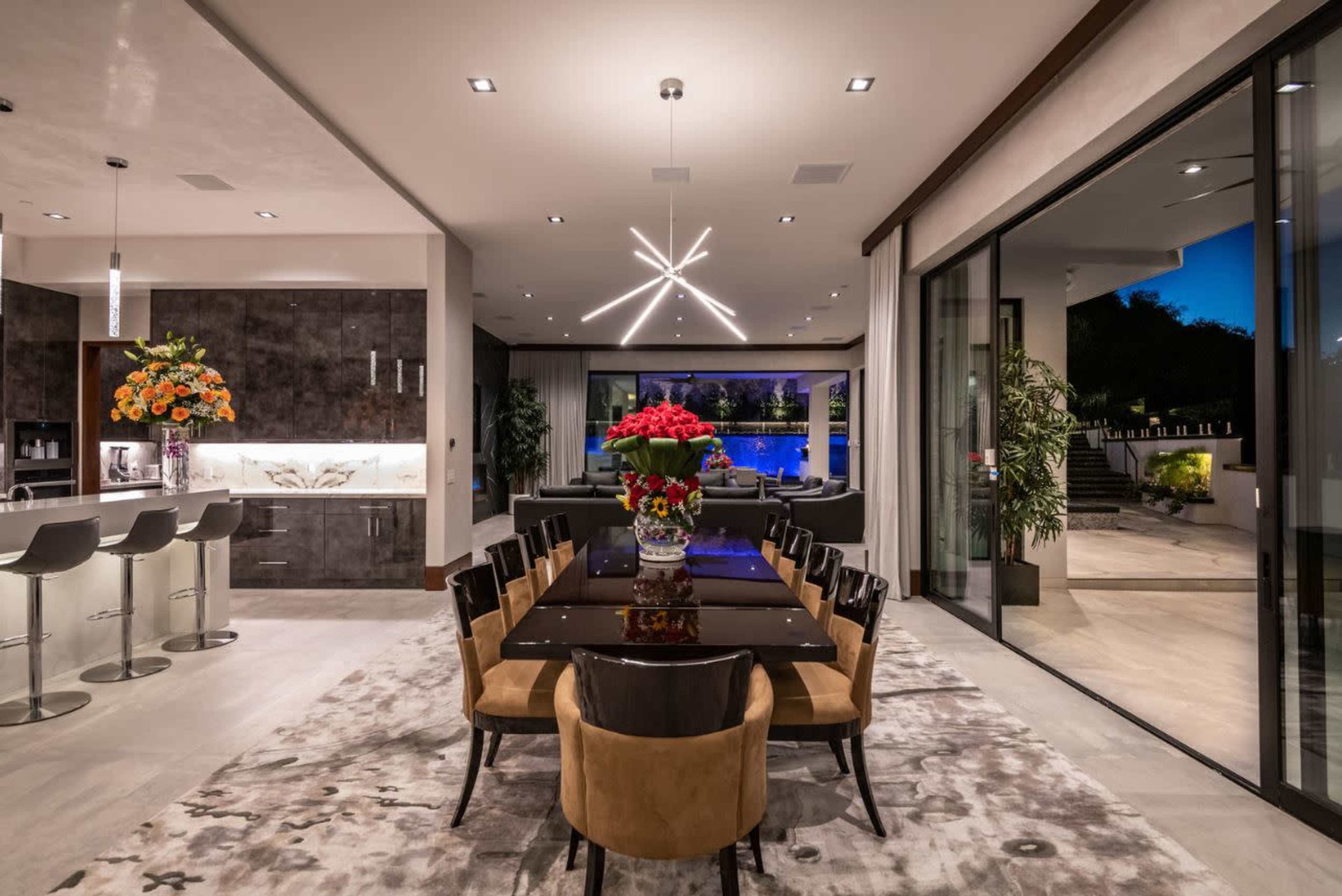 The image shows a modern dining area with a long black table surrounded by brown chairs, a large vase of flowers as a centerpiece, and views of a swimming pool through large glass doors.