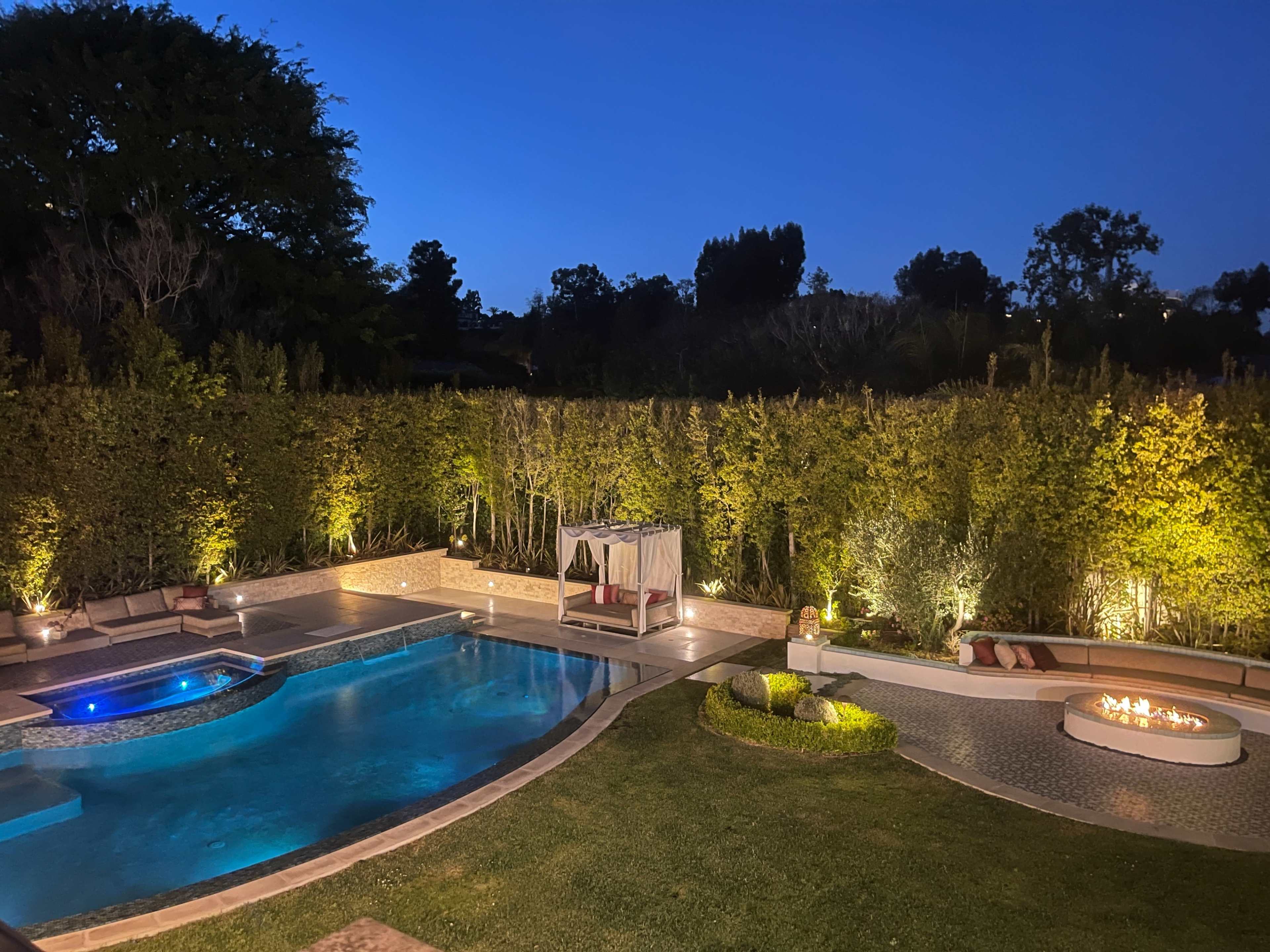 A well-lit backyard pool area surrounded by hedges, featuring a seating arrangement by the fire pit and a gazebo.