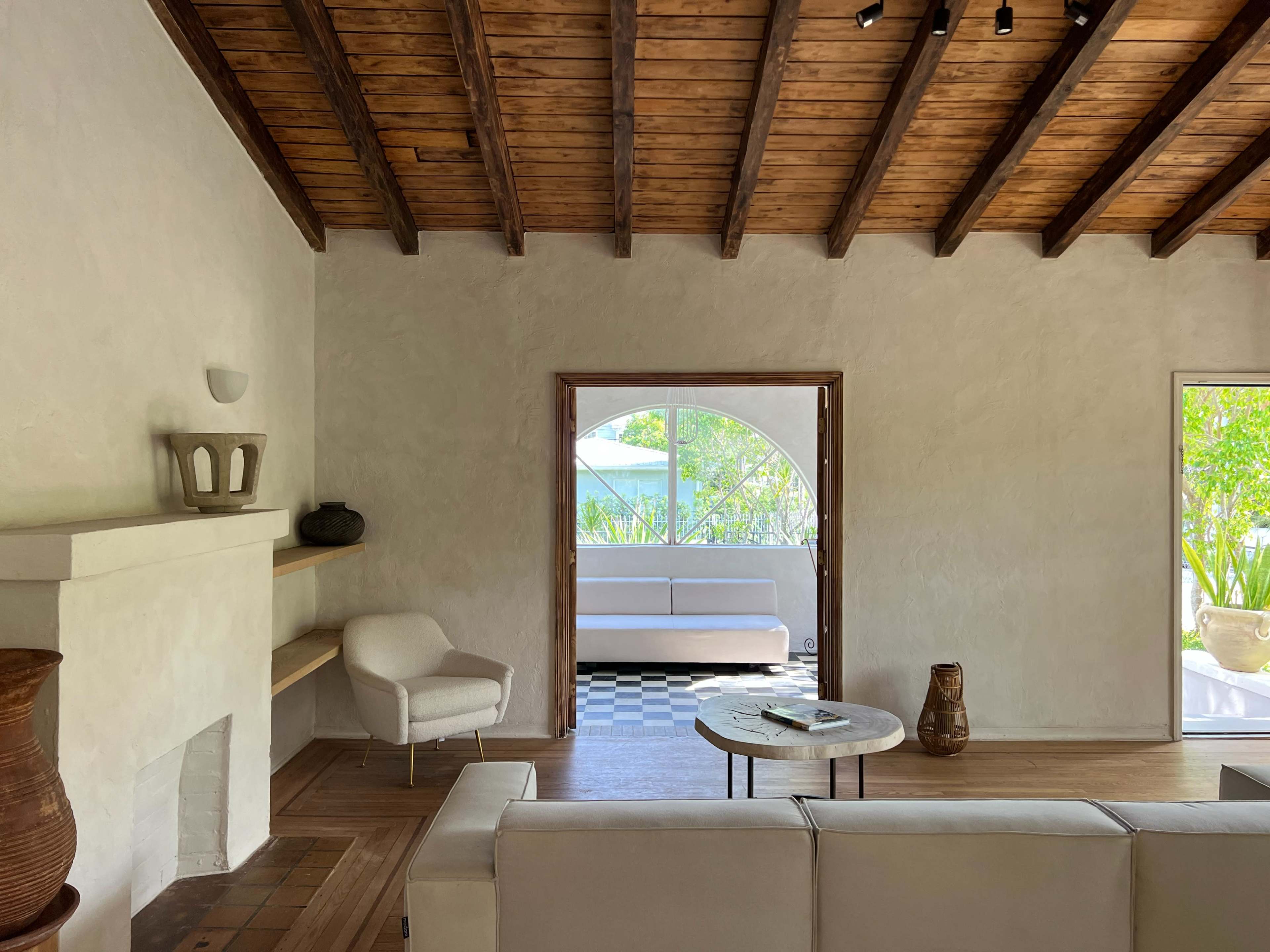 The image shows a spacious living room with a wooden ceiling, a modern white sofa, and large windows overlooking a green outdoor area.