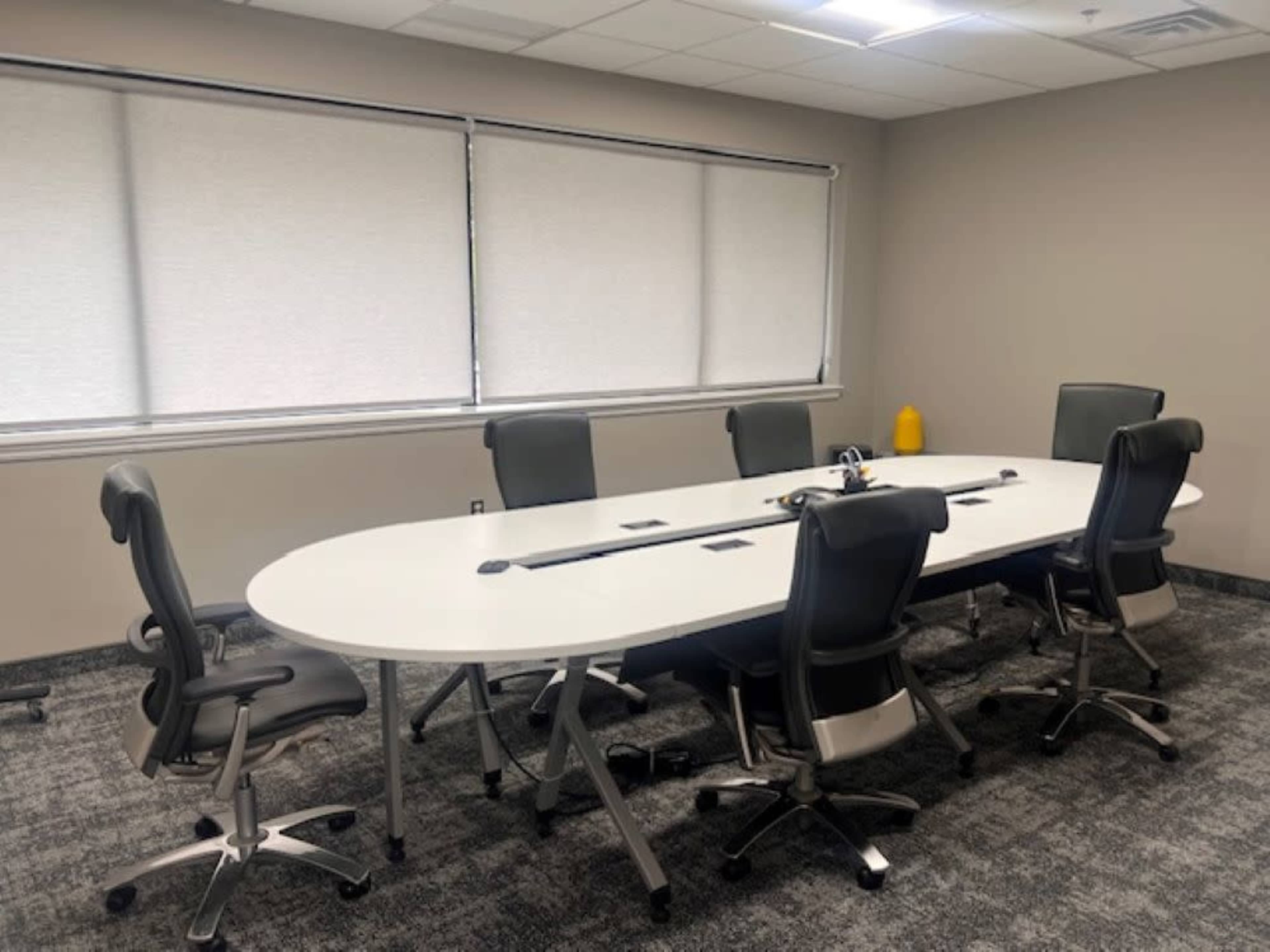 The image shows a modern conference room with an oval table surrounded by six ergonomic chairs and large windows covered by shades.