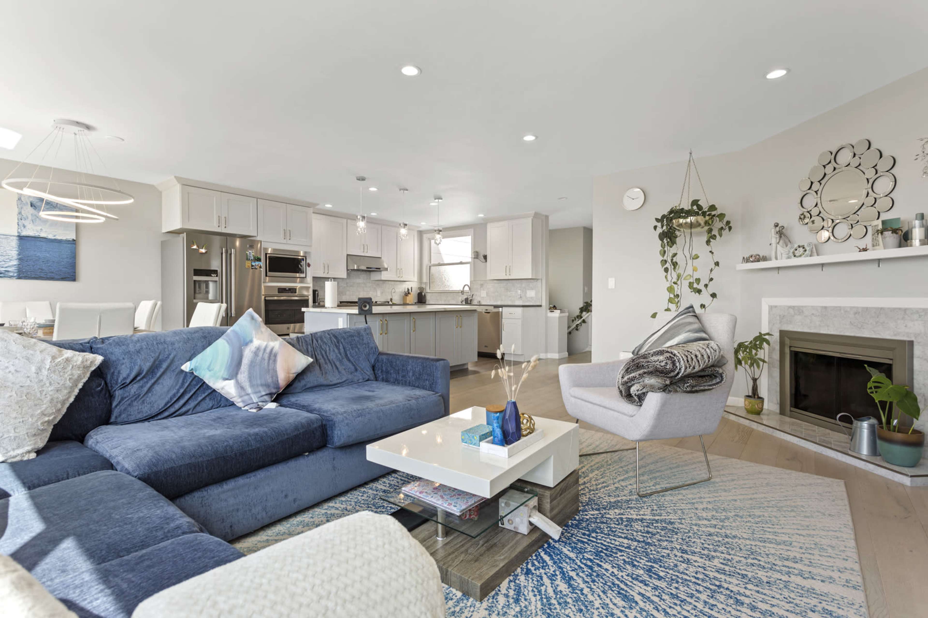 A modern living area with a blue sectional sofa, a coffee table, and an open kitchen in the background, complete with light cabinetry and a dining area.