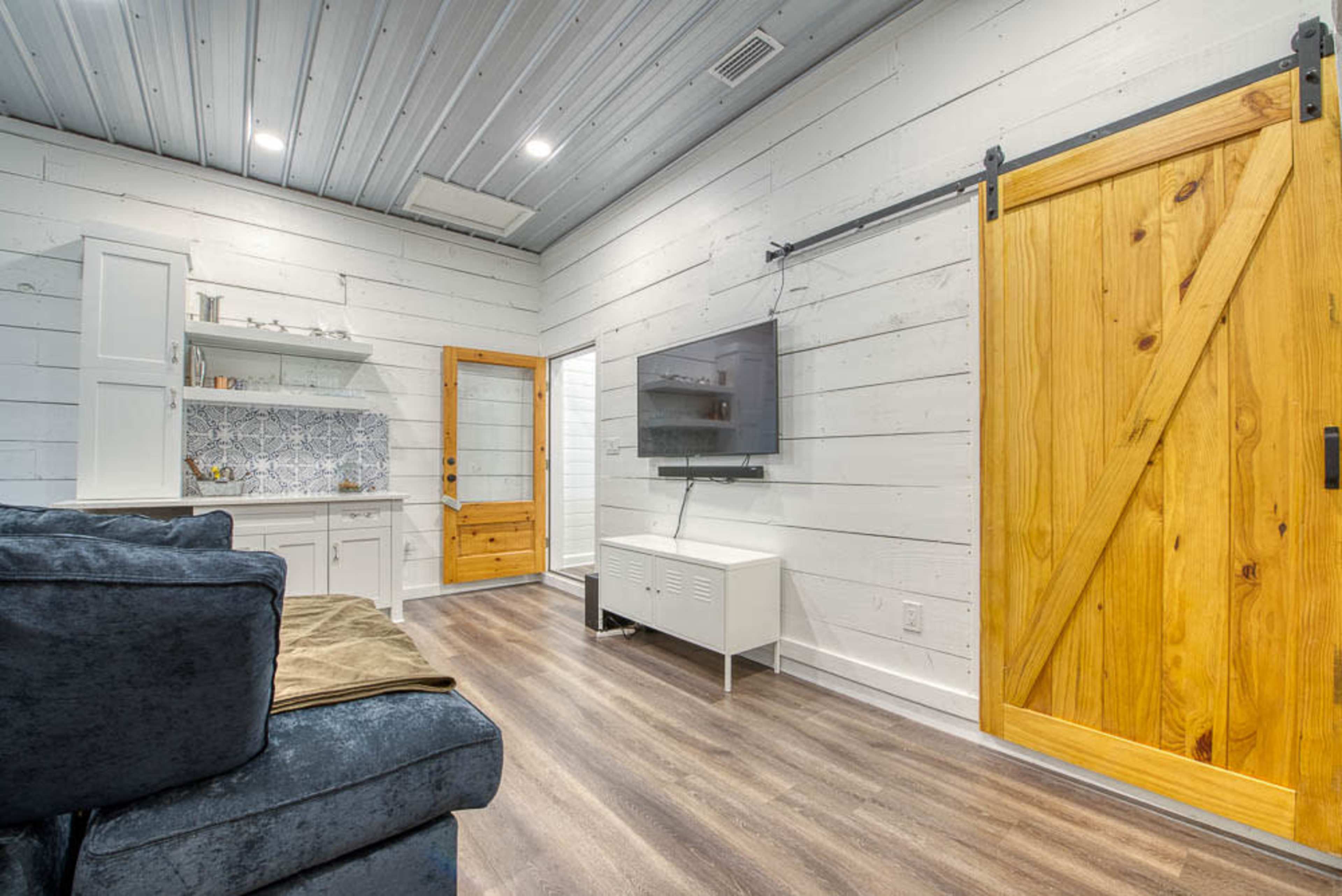 A modern interior living space featuring a sectional sofa, a wall-mounted television, a wooden barn door, and a small kitchenette area with white cabinets.